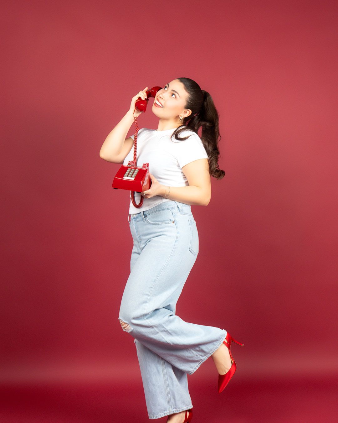 Young woman in white tee and wide-leg jeans poses playfully with a red rotary phone against a red background.