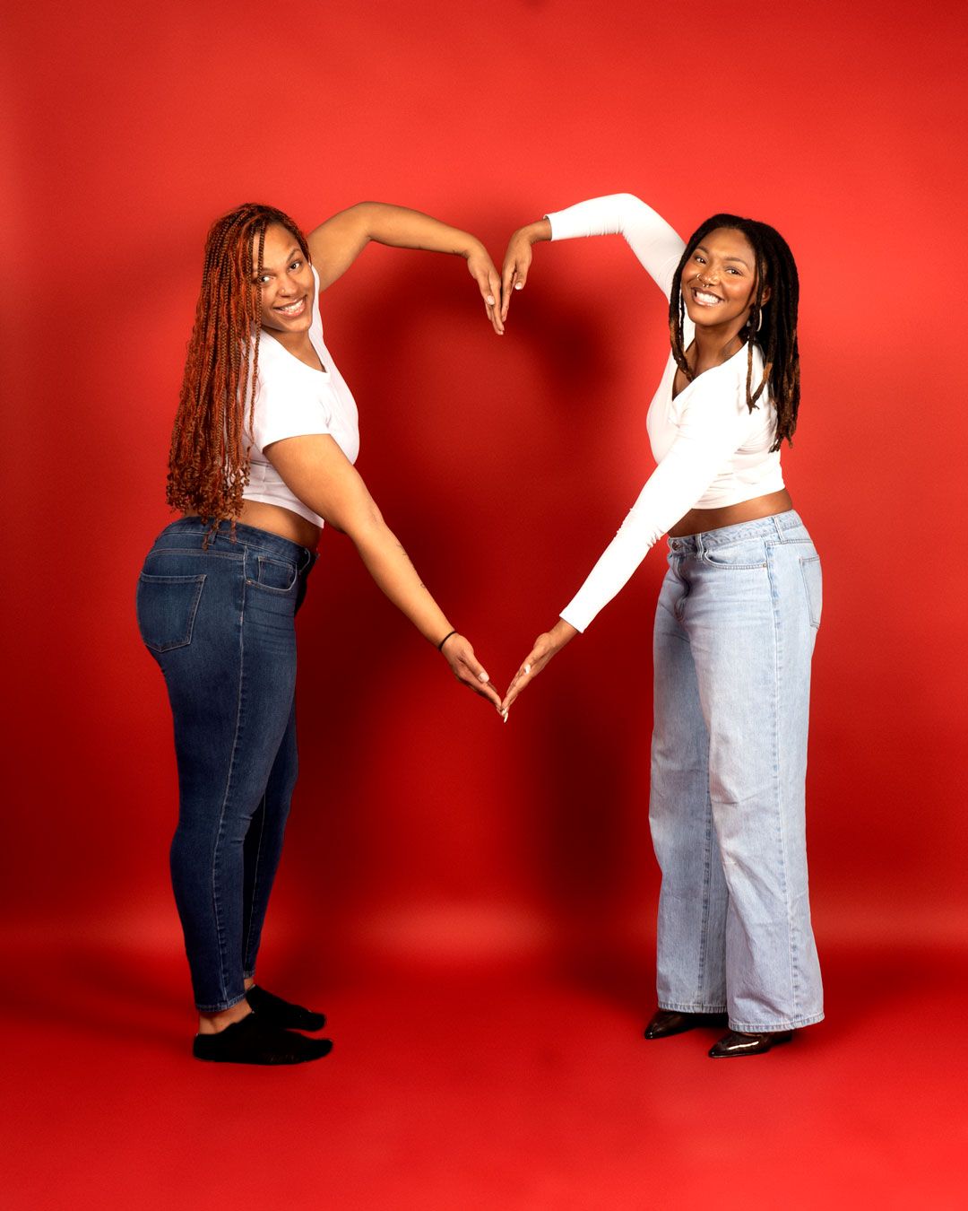 Two women in white tops and jeans form a heart shape with their arms against a red background.