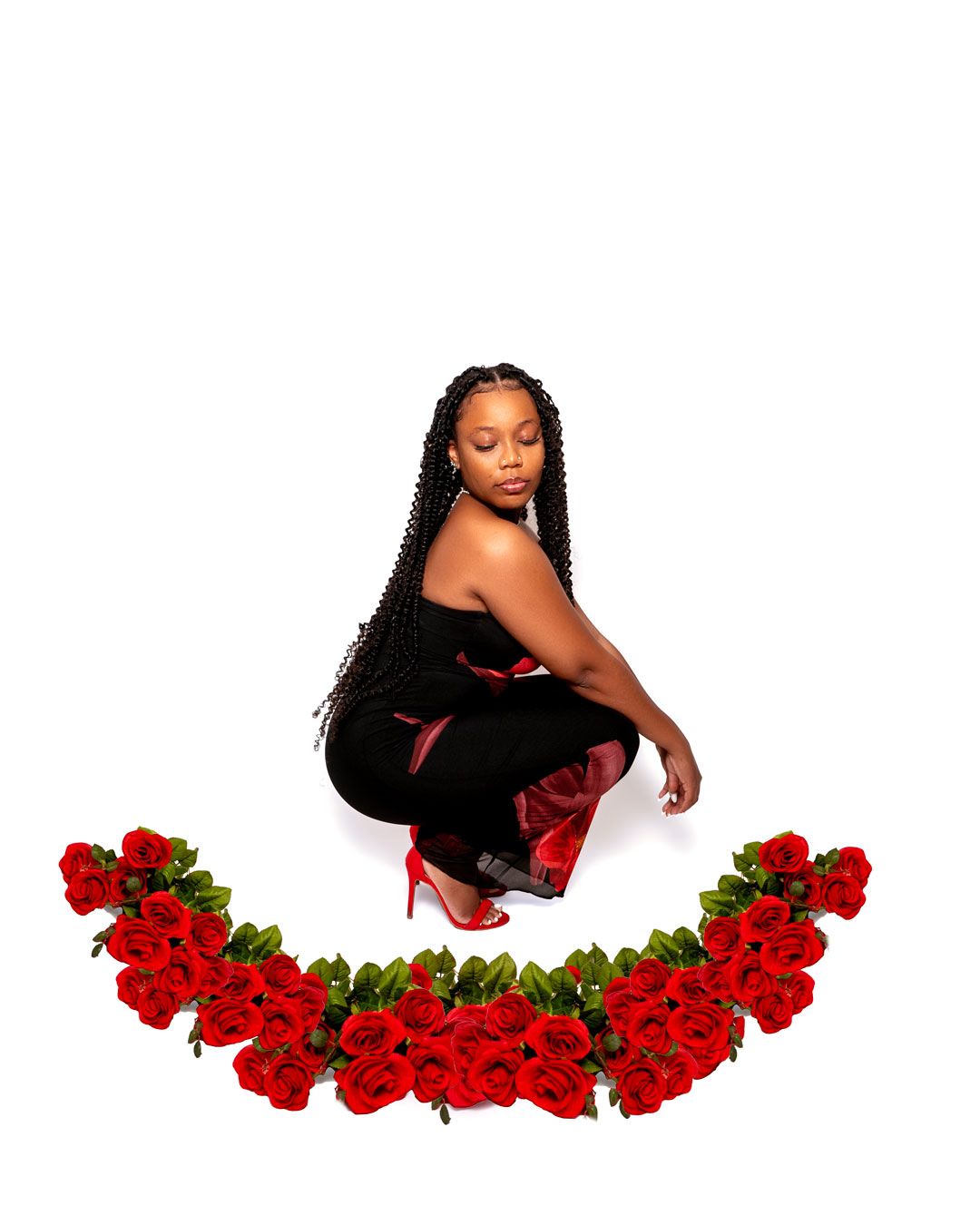 A person in a black outfit poses in a crouching position against a white backdrop with red roses arranged below.