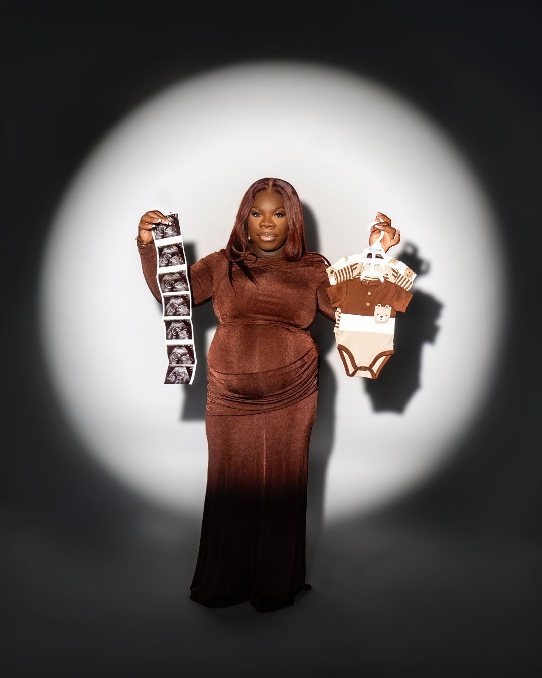 Pregnant woman in brown velvet gown holding ultrasound strip and baby onesie against spotlight background.