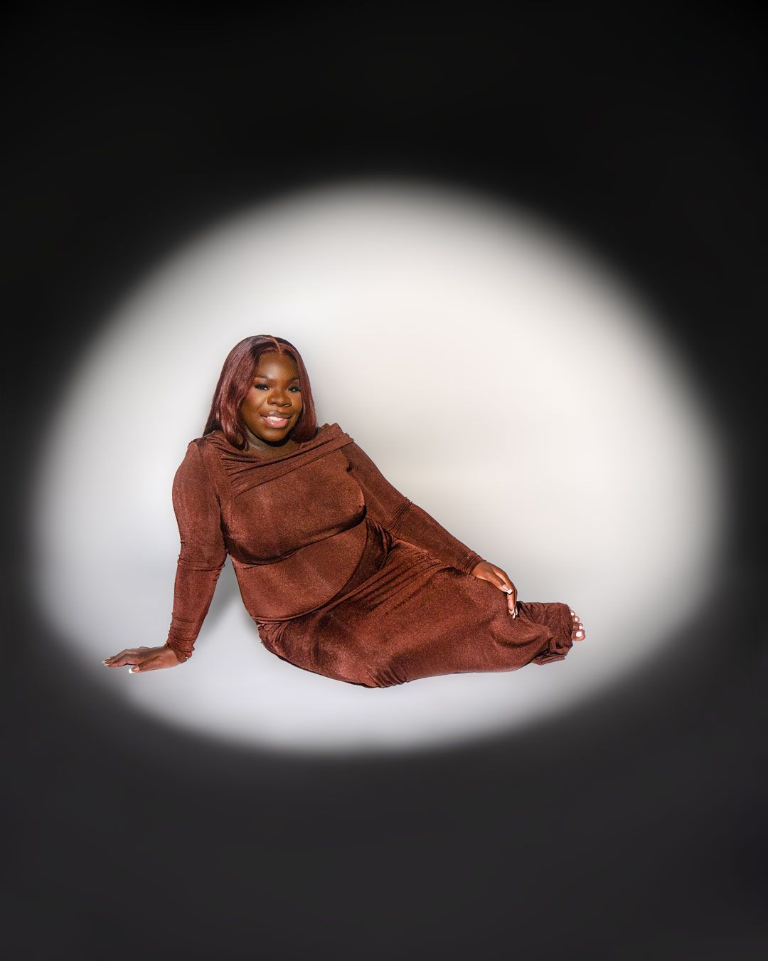 A woman in a brown outfit sits gracefully on the floor, illuminated by a spotlight against a dark background.