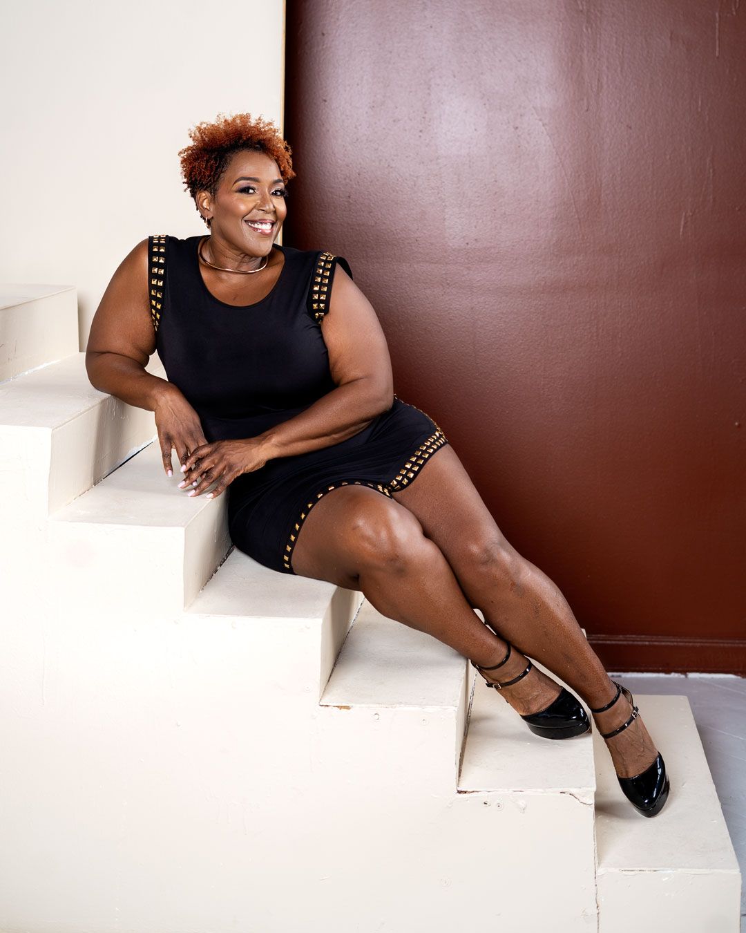 A smiling woman in a black studded mini dress sits on white stairs, posing confidently against a brown wall.