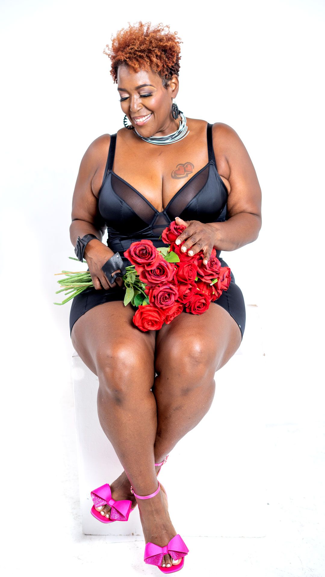 A plus-size woman in a black bodysuit and pink heels sits smiling while holding a bouquet of red roses.