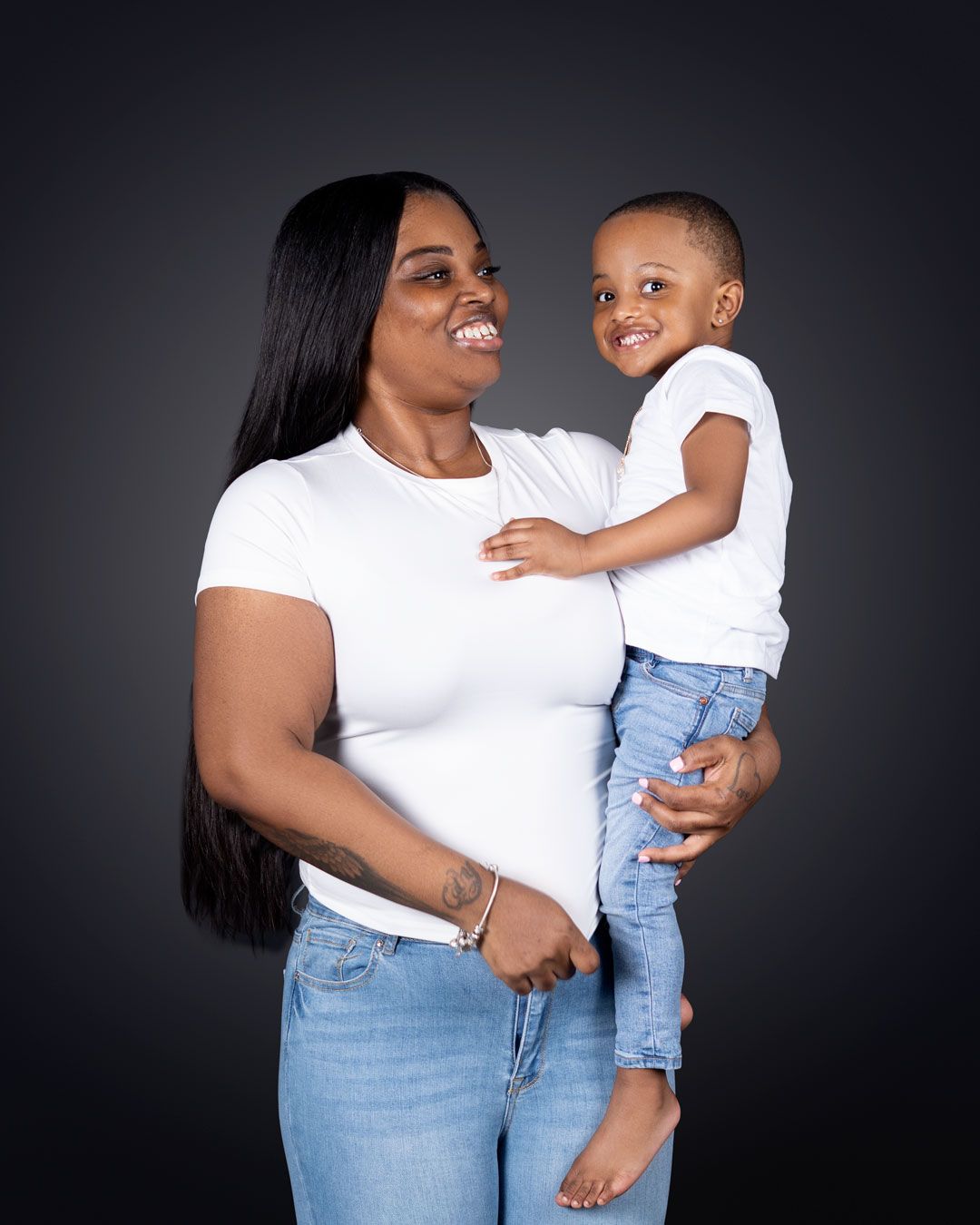 A smiling woman in white t-shirt and jeans holds a happy toddler boy against a dark studio background.