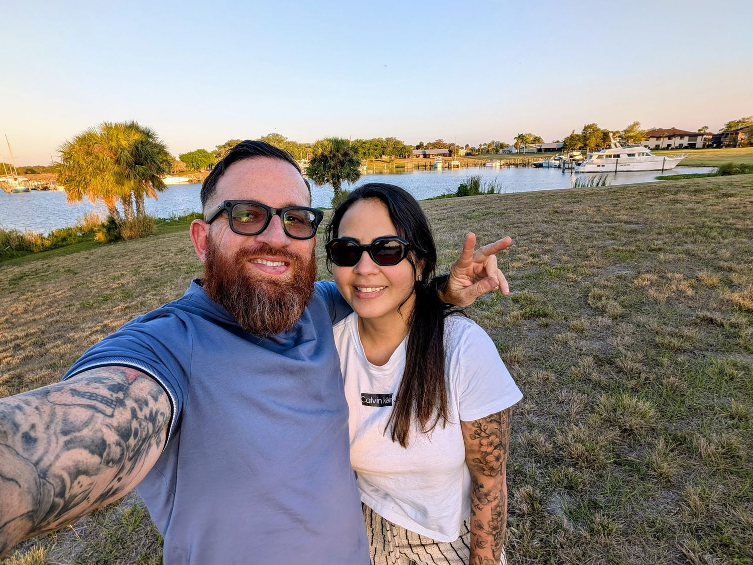 Couple takes selfie by waterfront during sunset with boats in background.