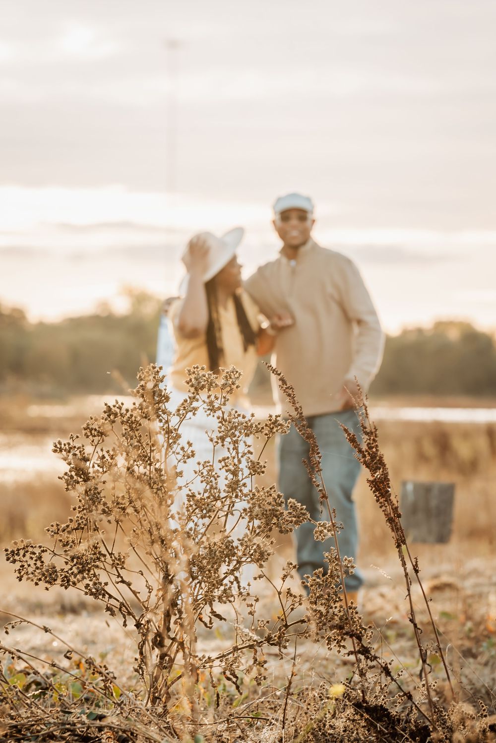 A couple stands in a golden field at sunset with dried wildflowers in the foreground.