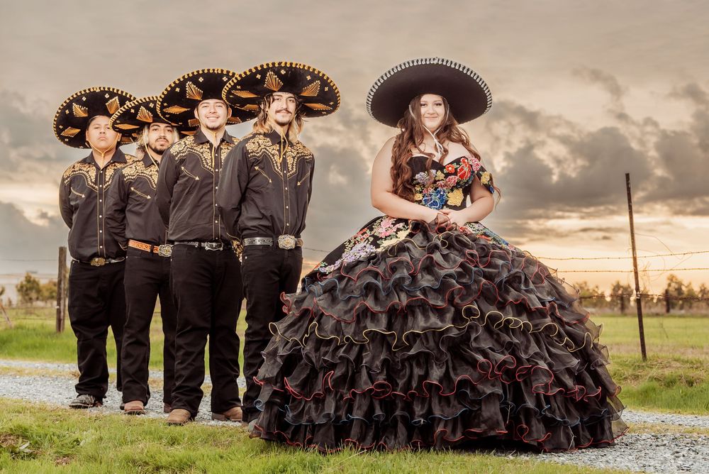 A group in black formal attire and traditional Mexican sombreros poses together outdoors at sunset for a quinceañera.