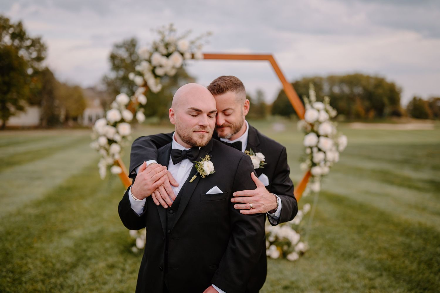 Two brides in formal attire stand together by a lake during their wedding photoshoot.