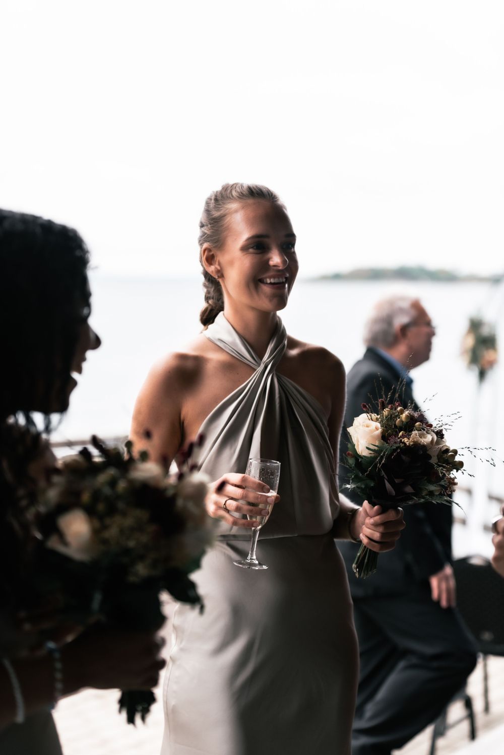 Elegant bridesmaid in halter neck dress holds champagne glass and bouquet while celebrating at waterfront wedding.