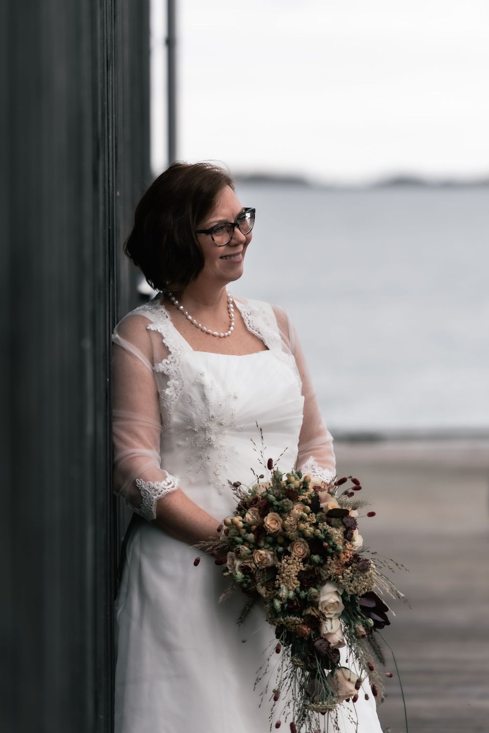 A bride in a white lace dress holds a romantic bouquet of flowers while standing near a waterfront.