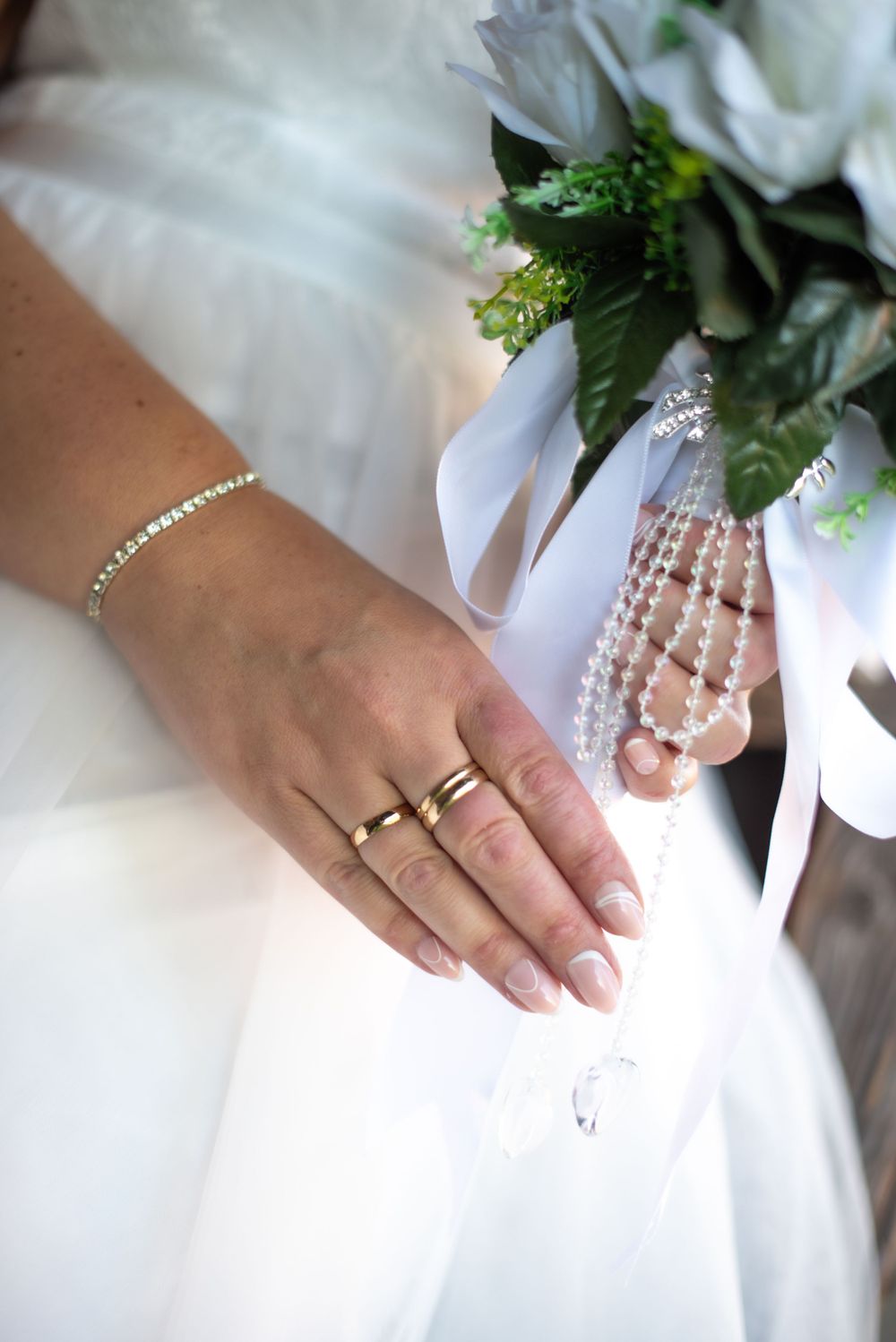 Close-up view of a bride's hand wearing wedding rings and bracelet with white dress and floral bouquet details.