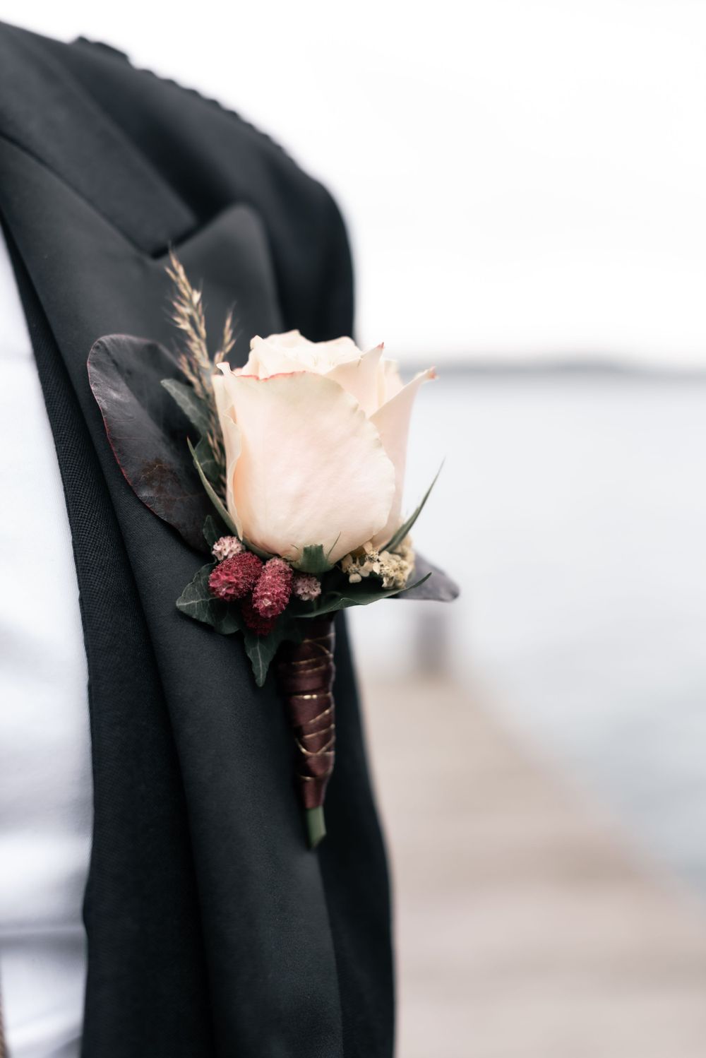 A cream-colored rose boutonniere with dark berries and greenery pinned against a black suit jacket.
