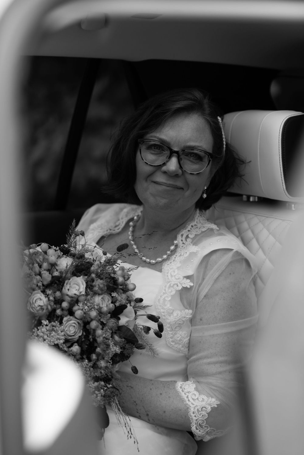 Black and white photo of a bride sitting in a car wearing glasses and holding a floral bouquet.