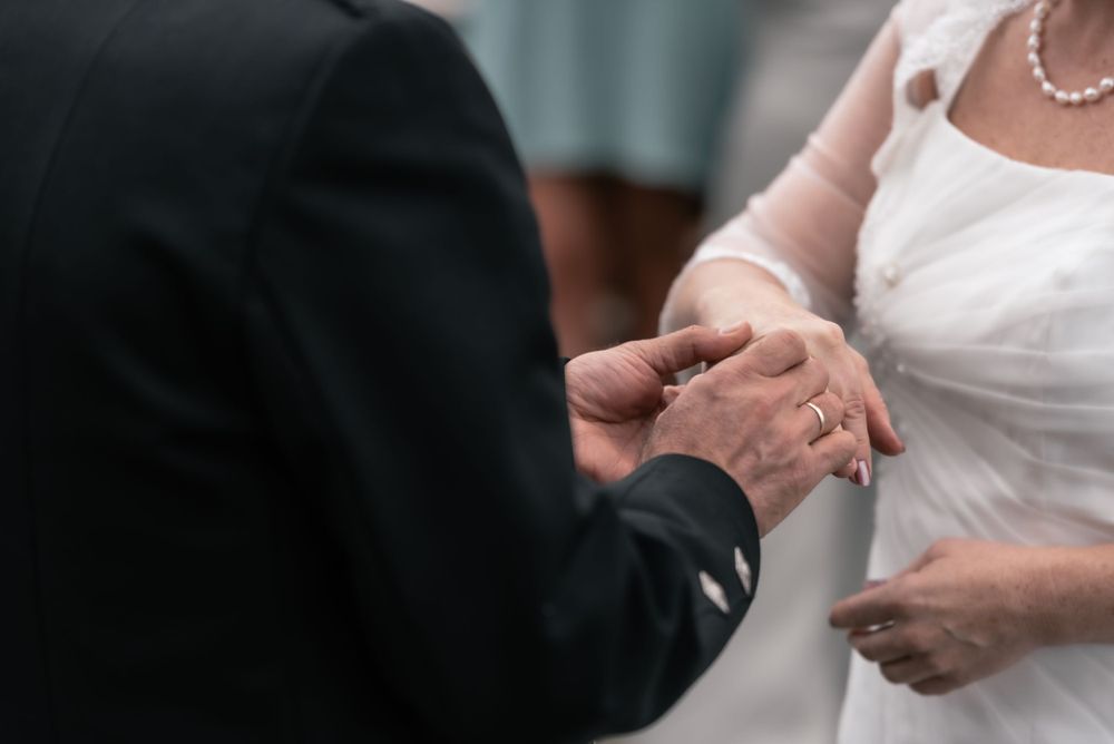 Close up of exchanging wedding rings during marriage ceremony with bride in white dress and groom in black suit.