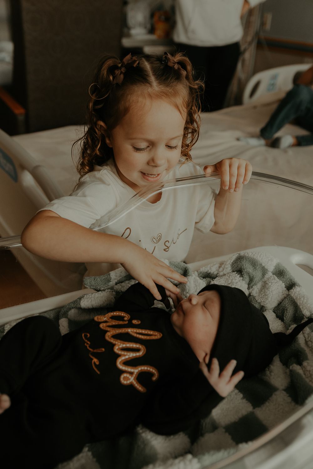 Child leaning over to observe newborn sibling in hospital bed.