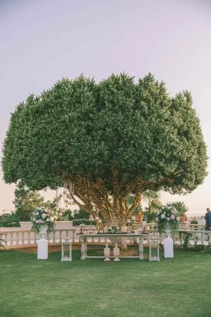 Elegant outdoor wedding setup under a large green tree with white floral arrangements on pedestals at dusk.