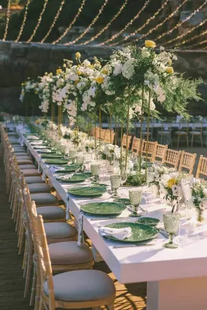 Elegant outdoor wedding reception with long white table, green charger plates, tall floral centerpieces, and gold chiavari chairs.