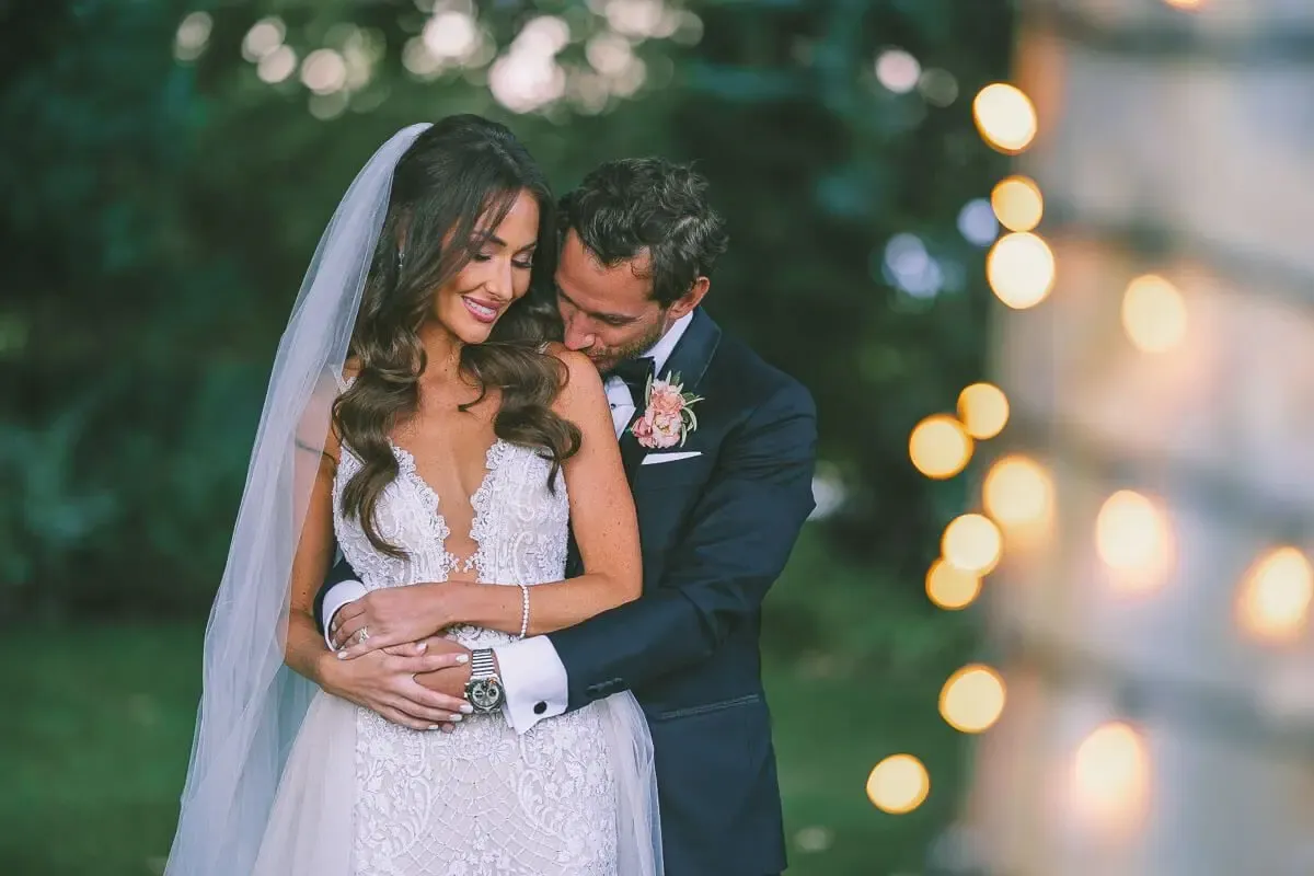 A bride and groom share a romantic embrace outdoors at twilight, with soft bokeh lights glowing in the background.