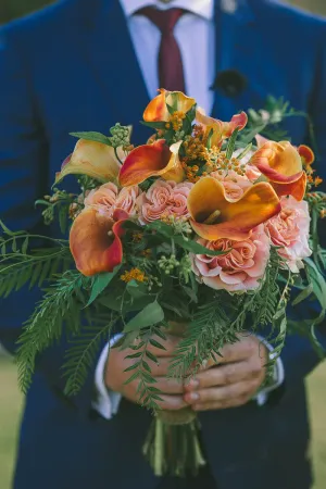 Man in navy suit holding vibrant orange calla lily and pink rose wedding bouquet with lush greenery.