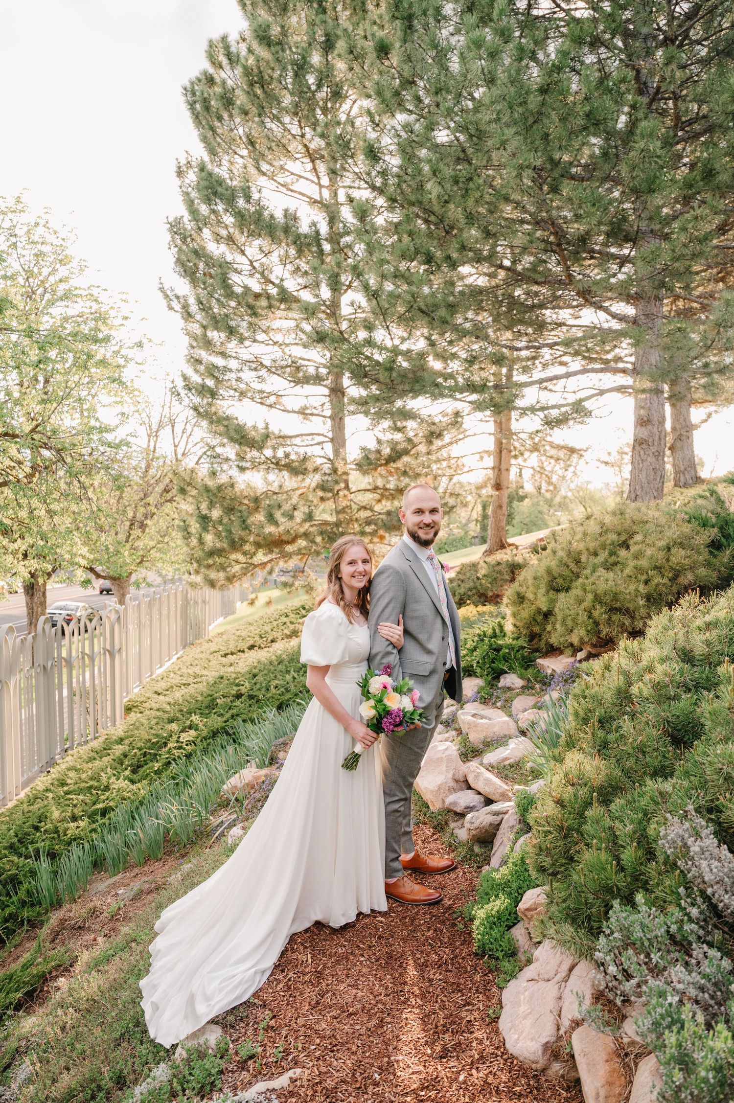A couple poses together on a mulch path in a garden surrounded by evergreen trees and flowering shrubs.