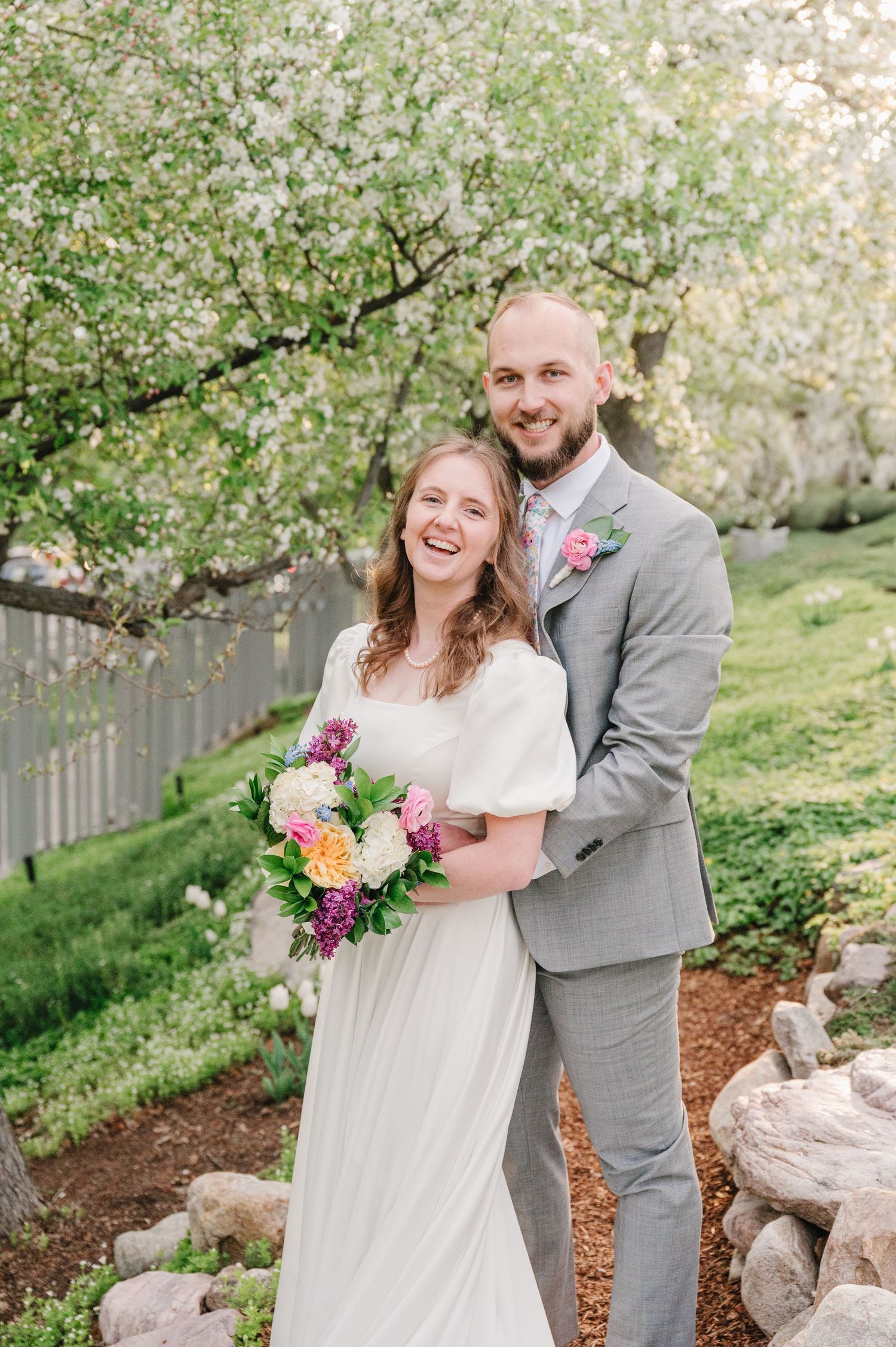 A couple stands together in a spring garden with blooming white cherry blossoms as a romantic backdrop.