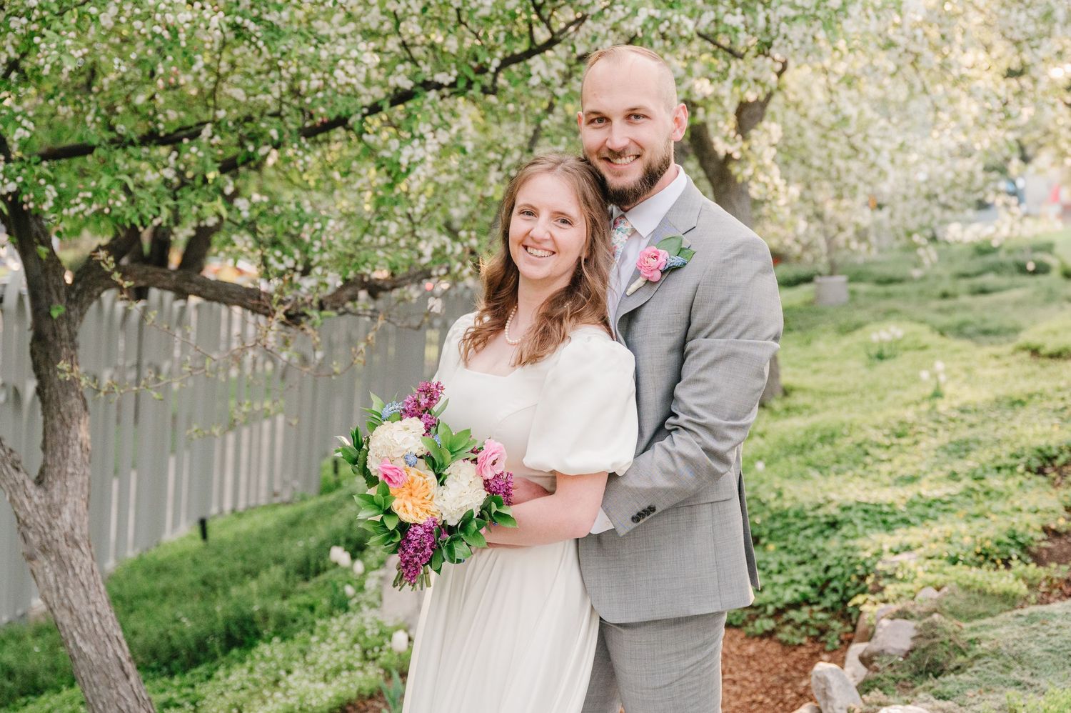 A couple poses outdoors in wedding attire with a colorful bouquet in front of a rustic fence and blooming spring trees.
