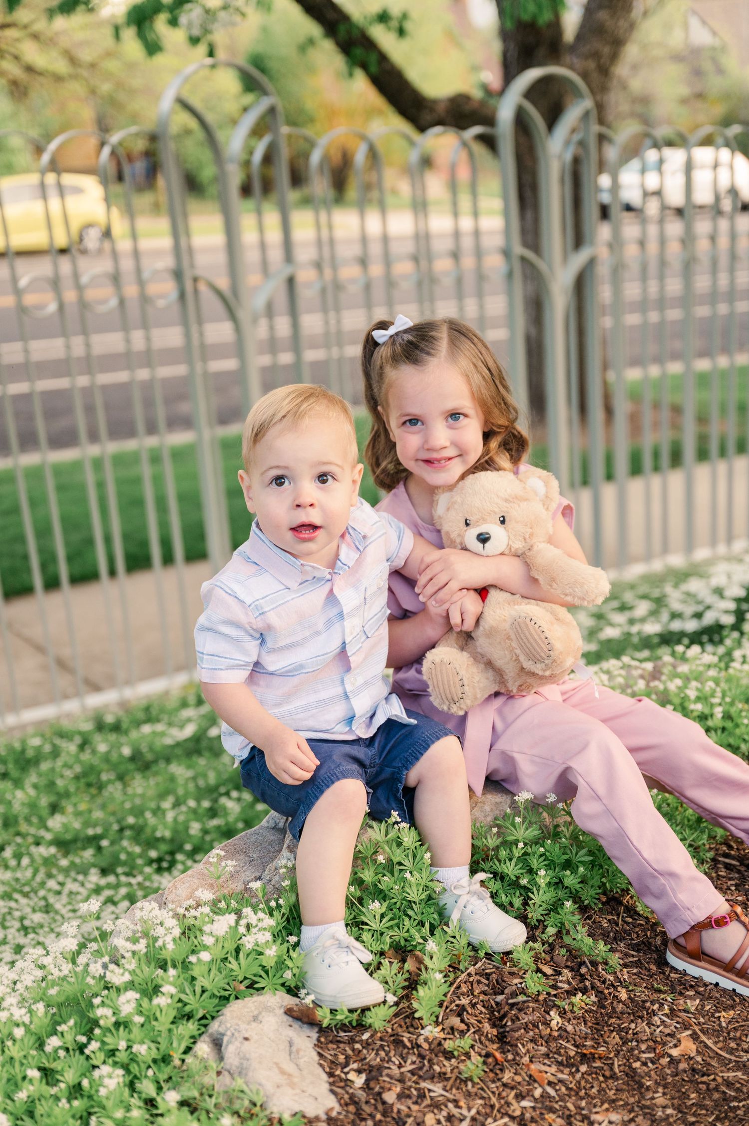 Two children sitting on garden rocks with teddy bear and ornamental fence in background.