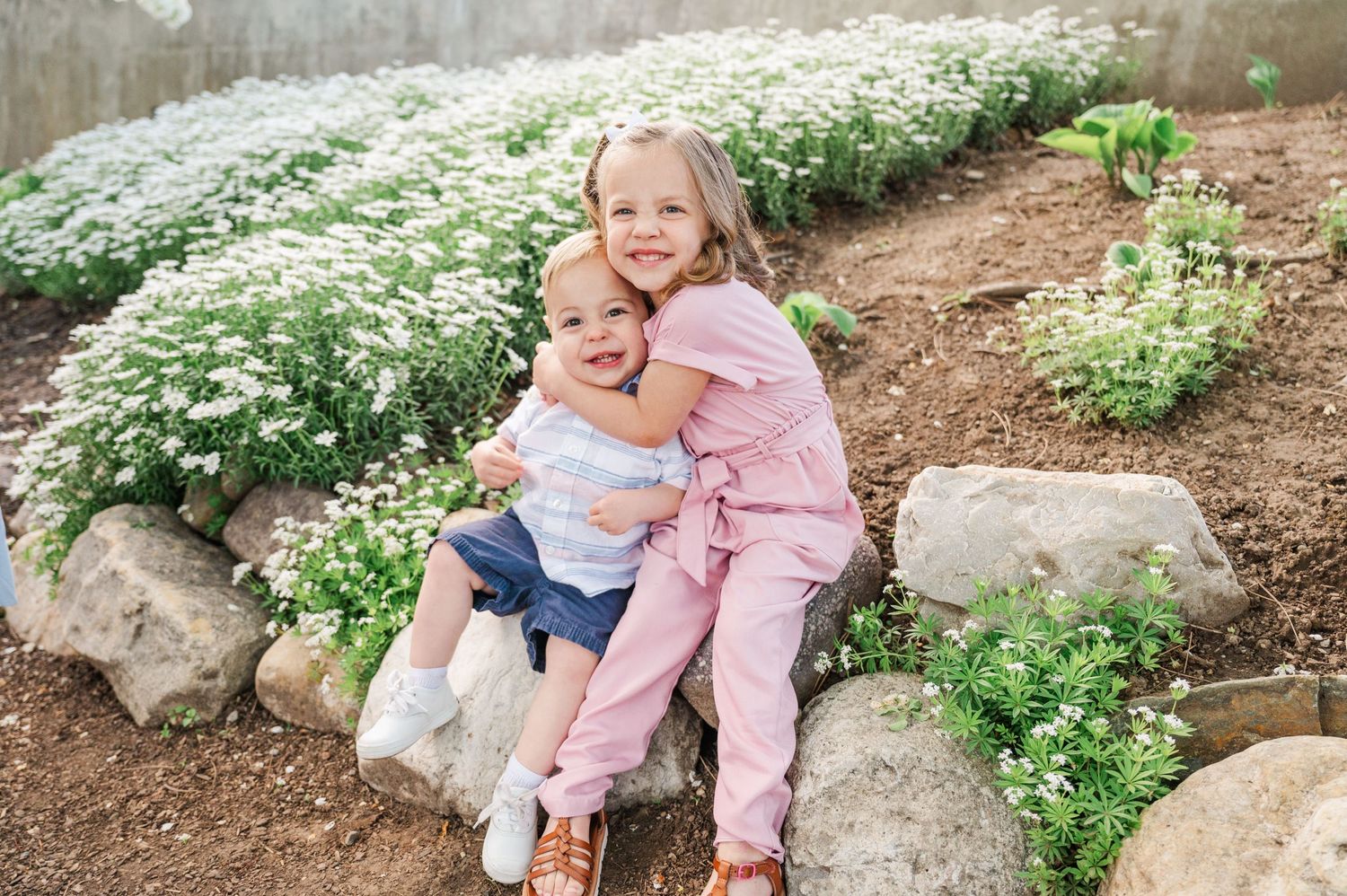 Two children hugging on garden rocks with white flowering plants in background.