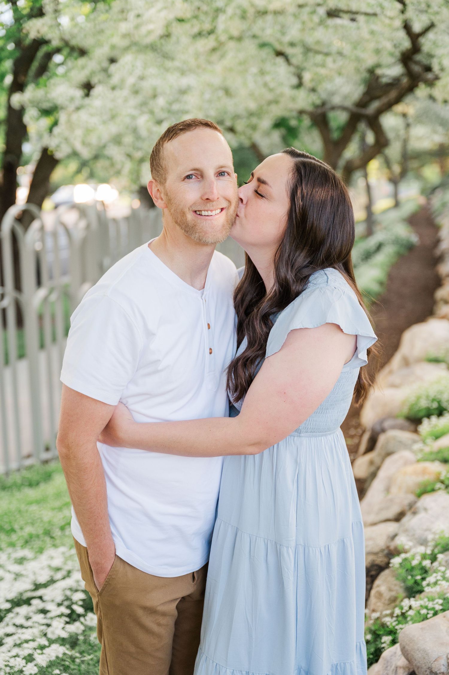 Romantic couple sharing tender moment in garden with blooming trees and white fence.