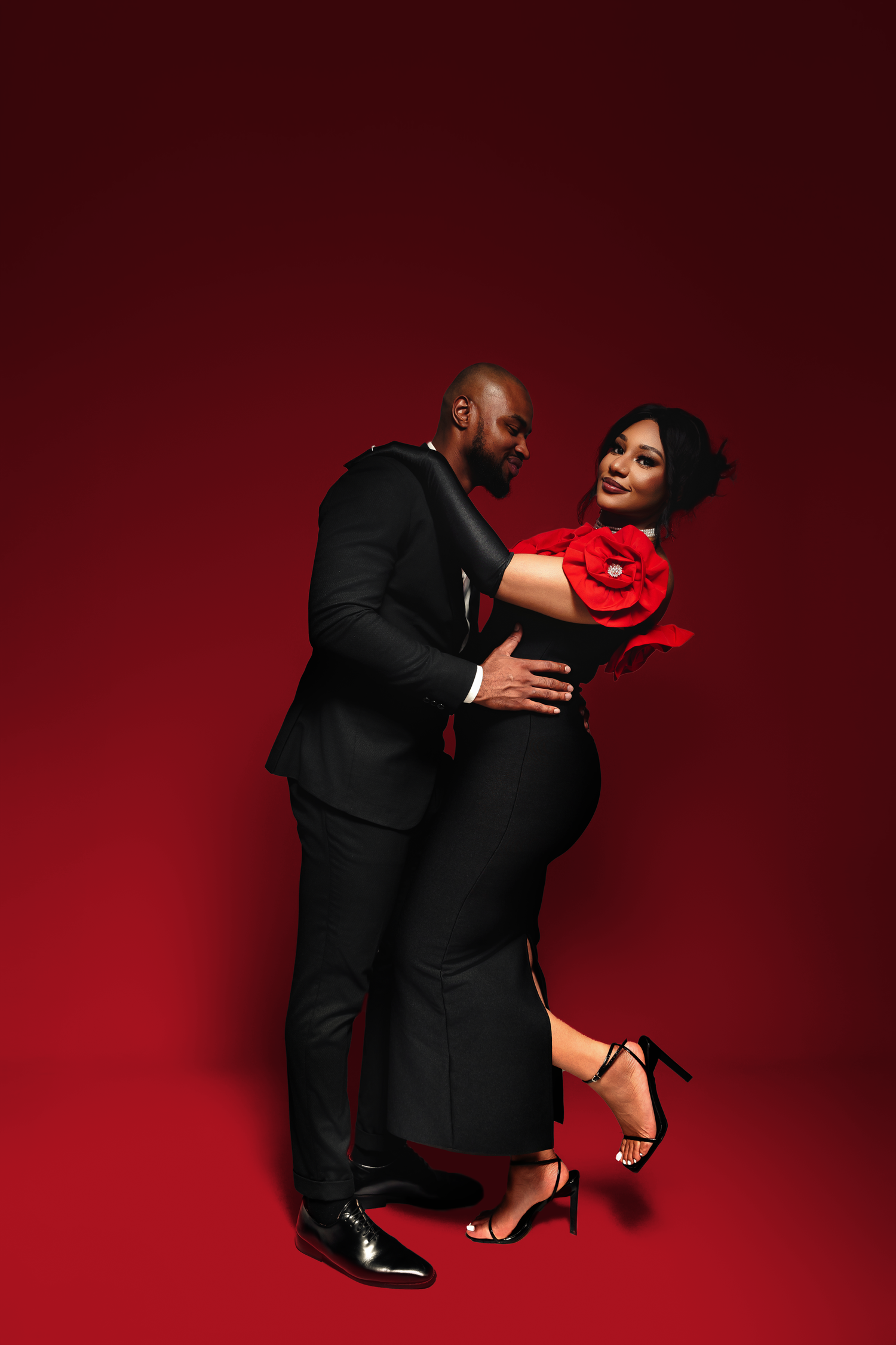 Elegant couple in black attire embrace romantically against a deep red backdrop with woman holding a red rose.