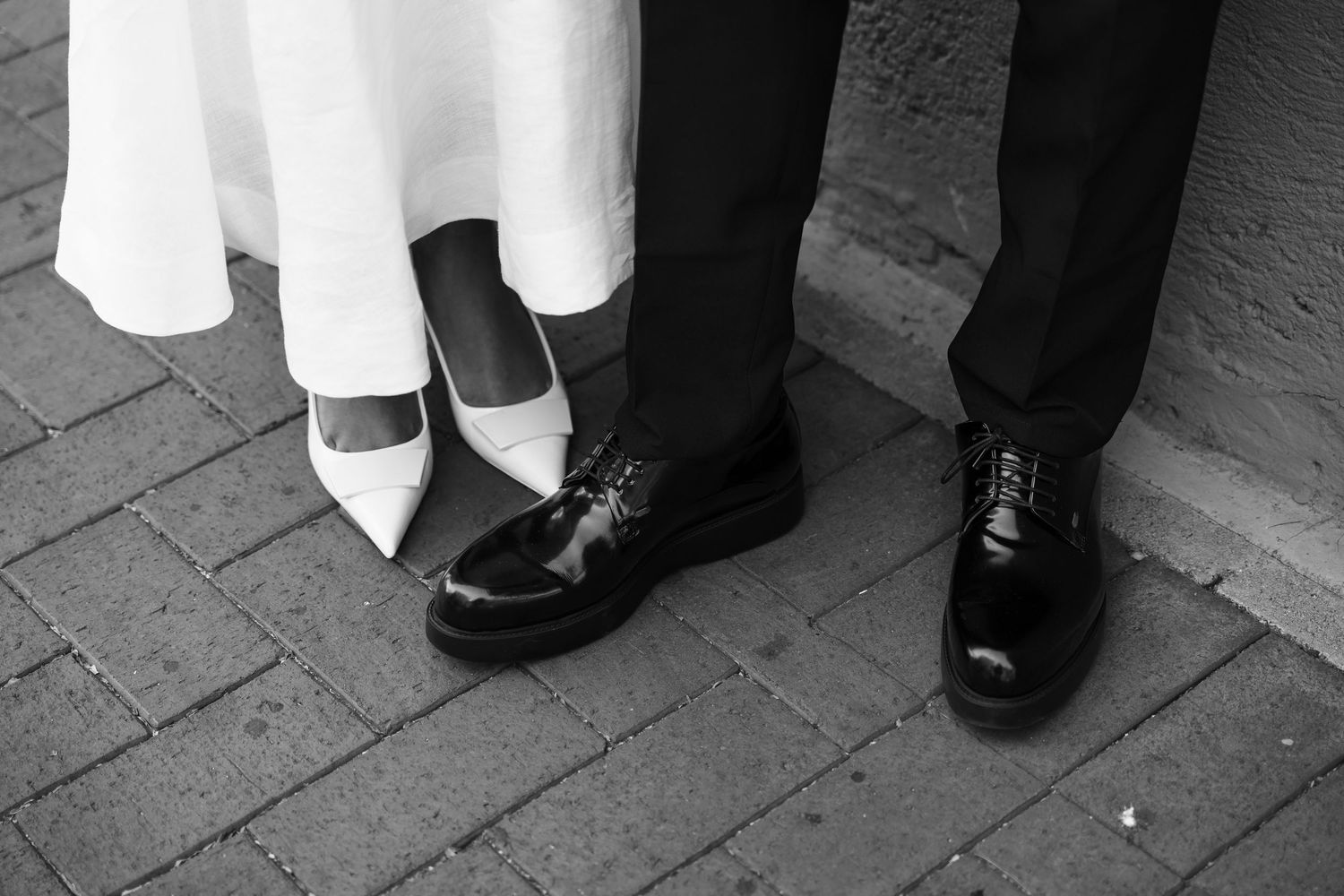 Black and white photograph shows formal dress shoes and white heels standing on a brick walkway.