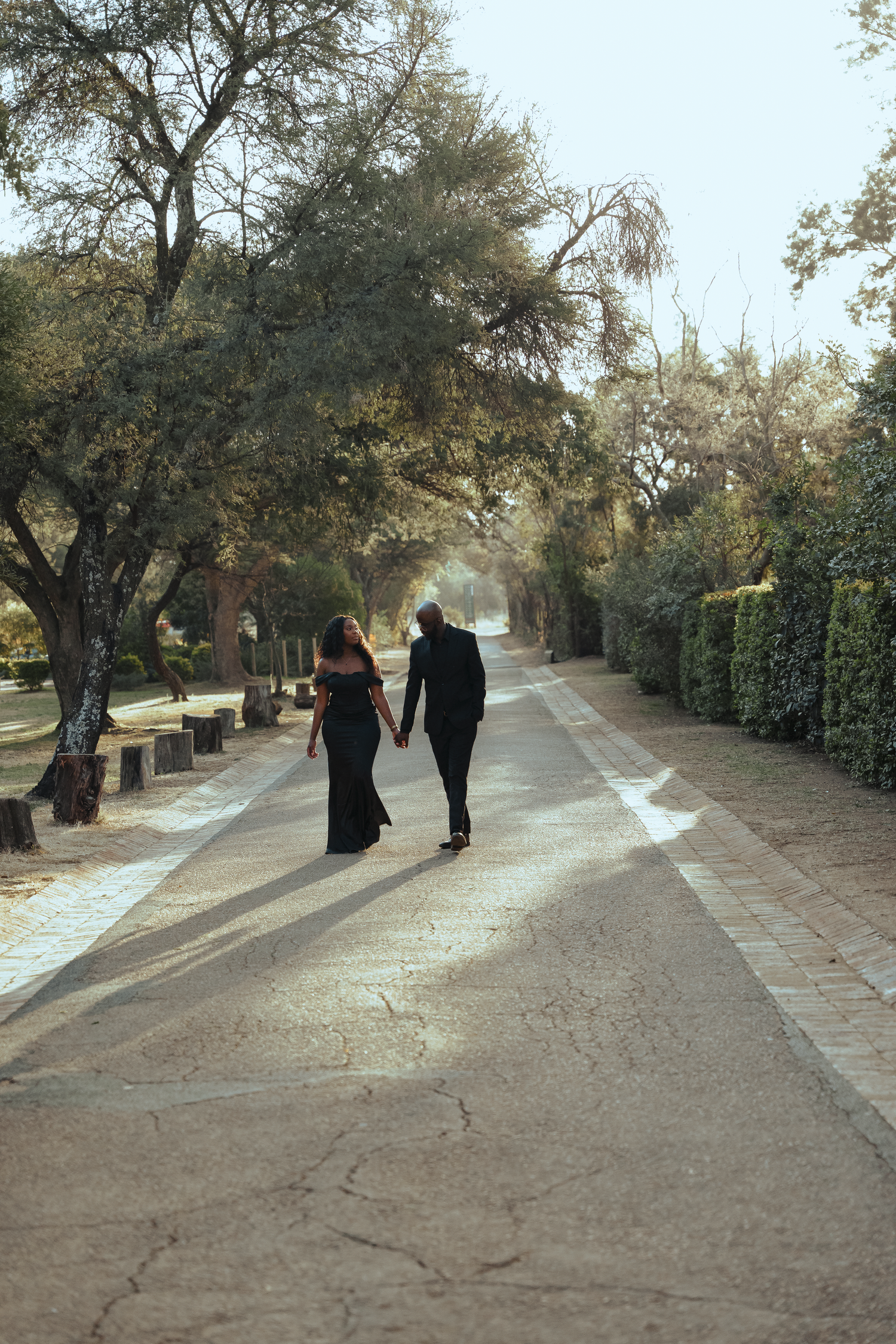 A couple in black attire walks together down a tree-lined path on a sunny day.