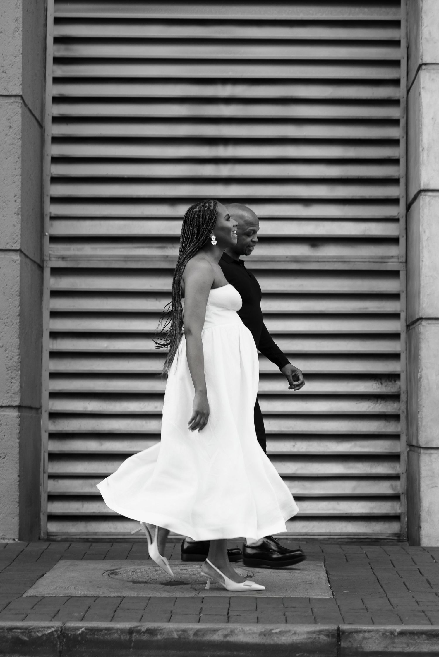 Black and white portrait of person in flowing white dress walking sideways against industrial metal backdrop.