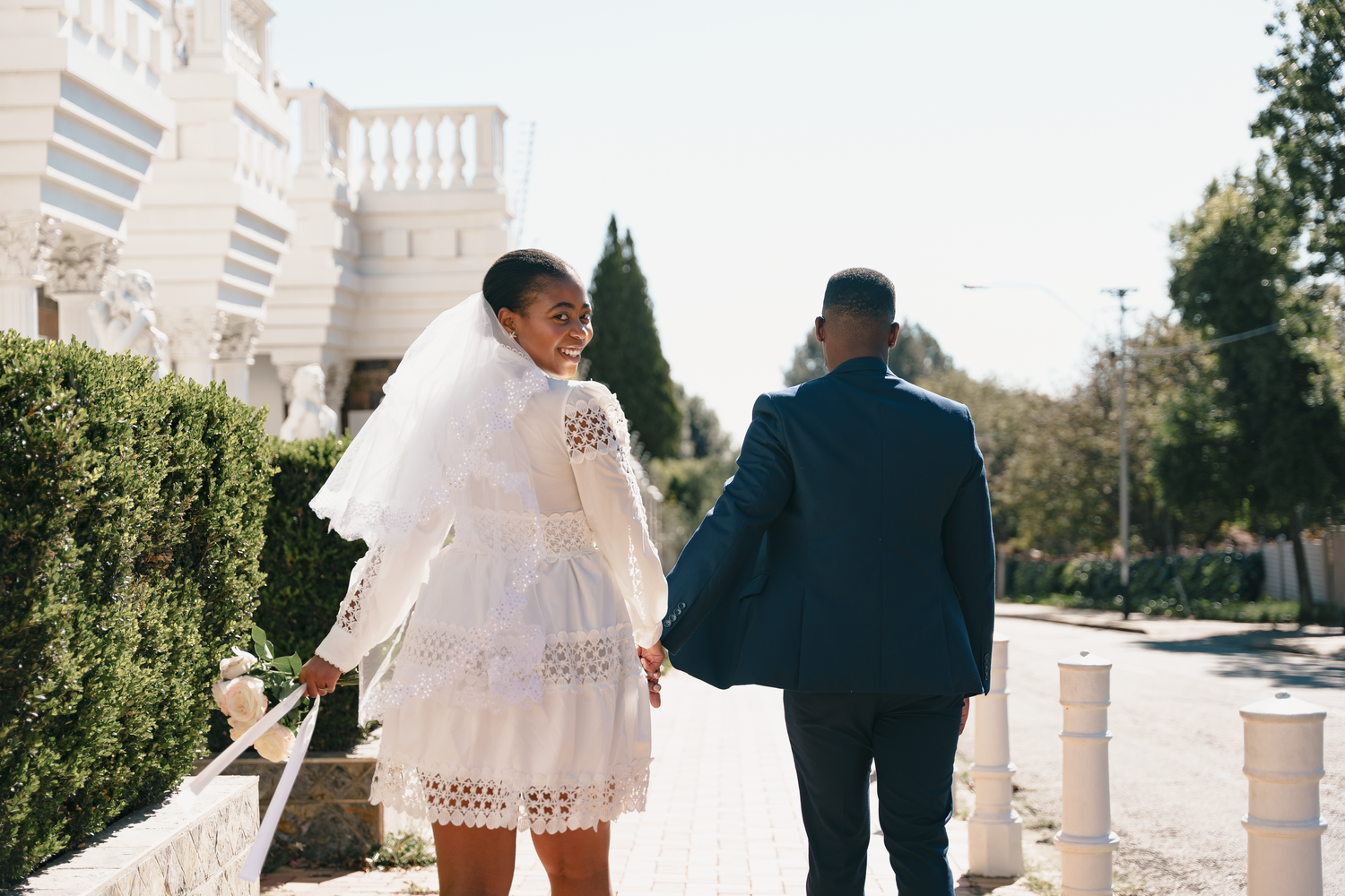 Couple walking outdoors, bride wearing short white lace dress and groom in dark suit against architectural backdrop.