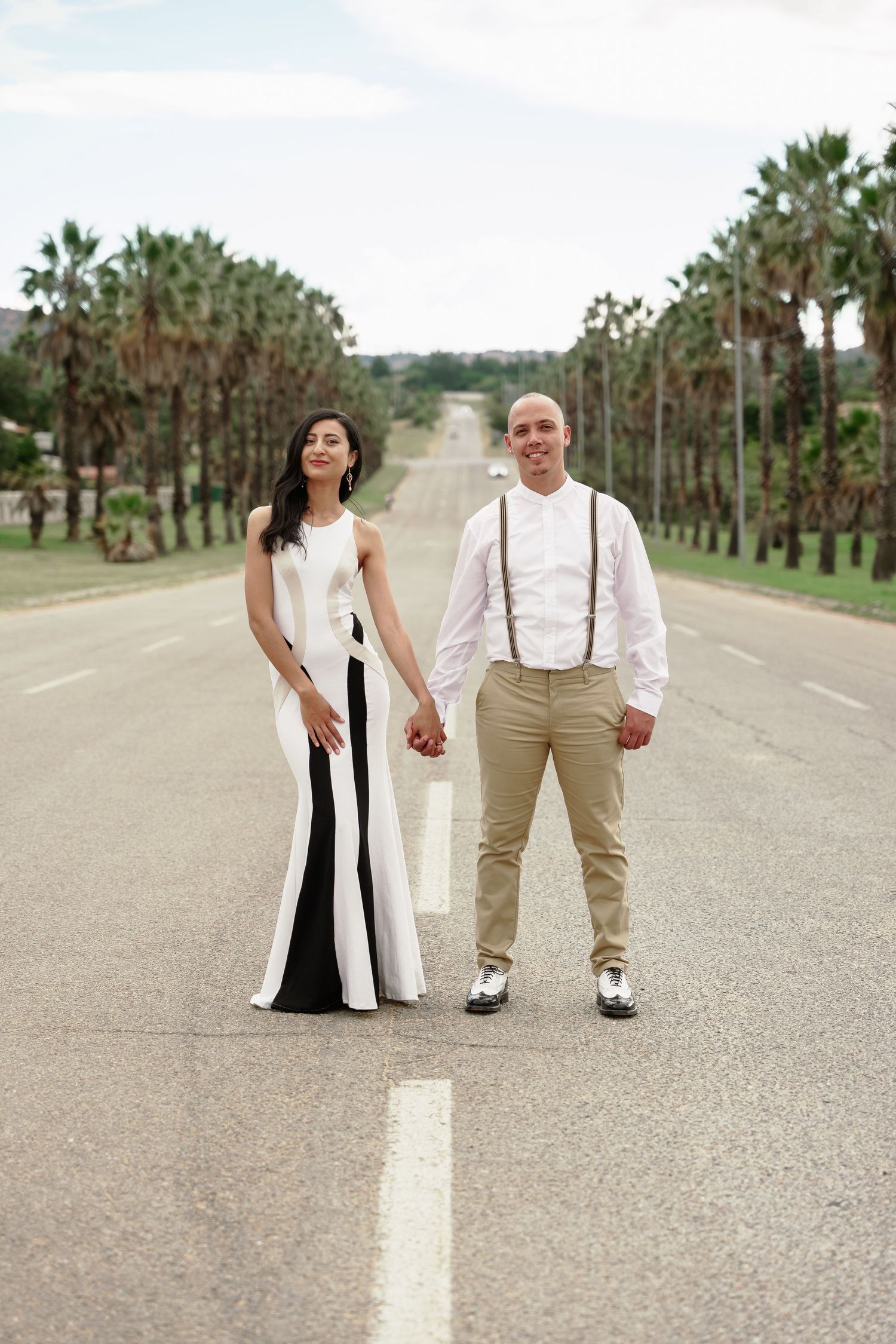 Stylish couple poses on palm tree-lined road in formal attire.