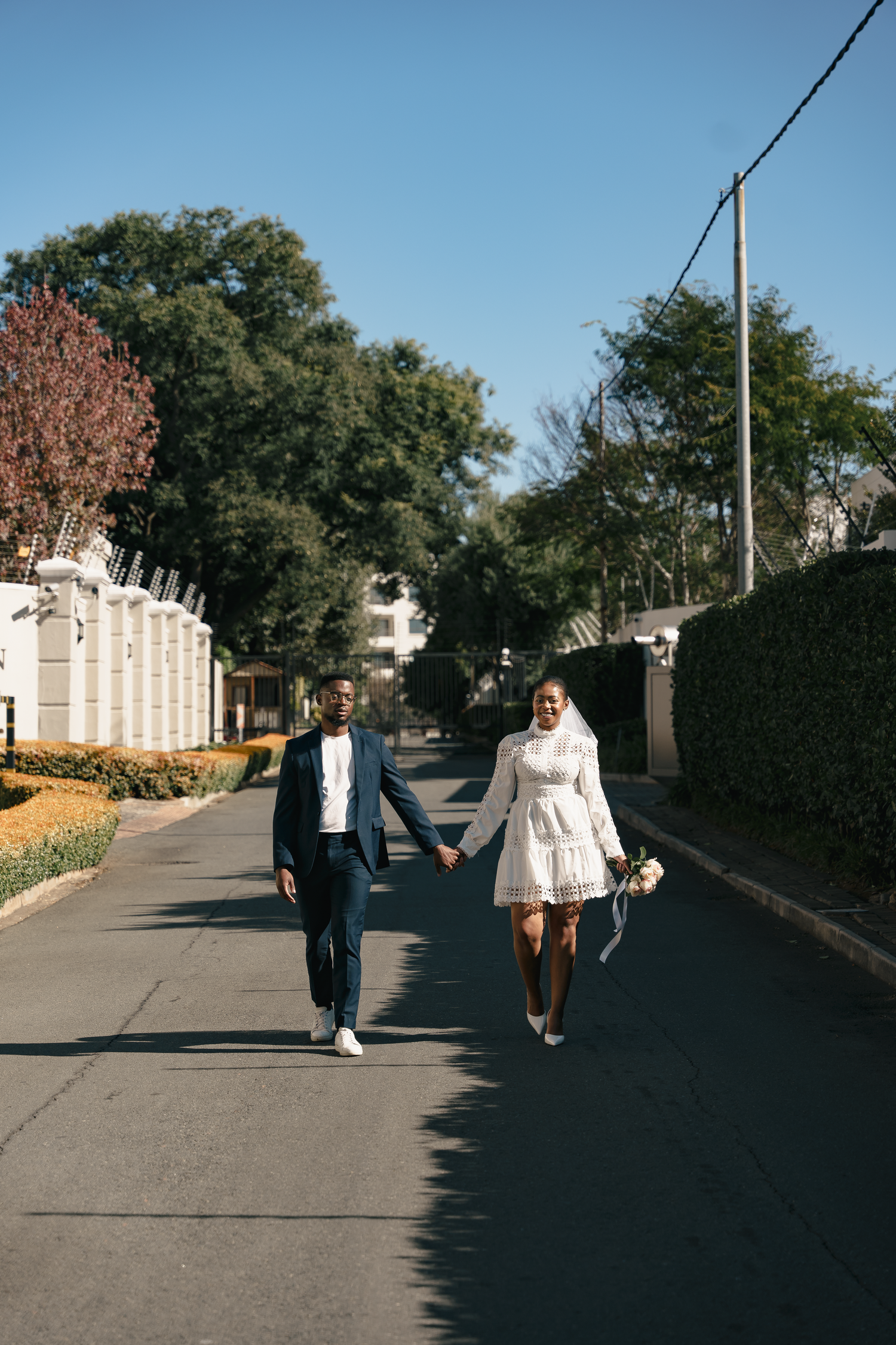 Couple holding hands while walking down tree-lined street in wedding attire.