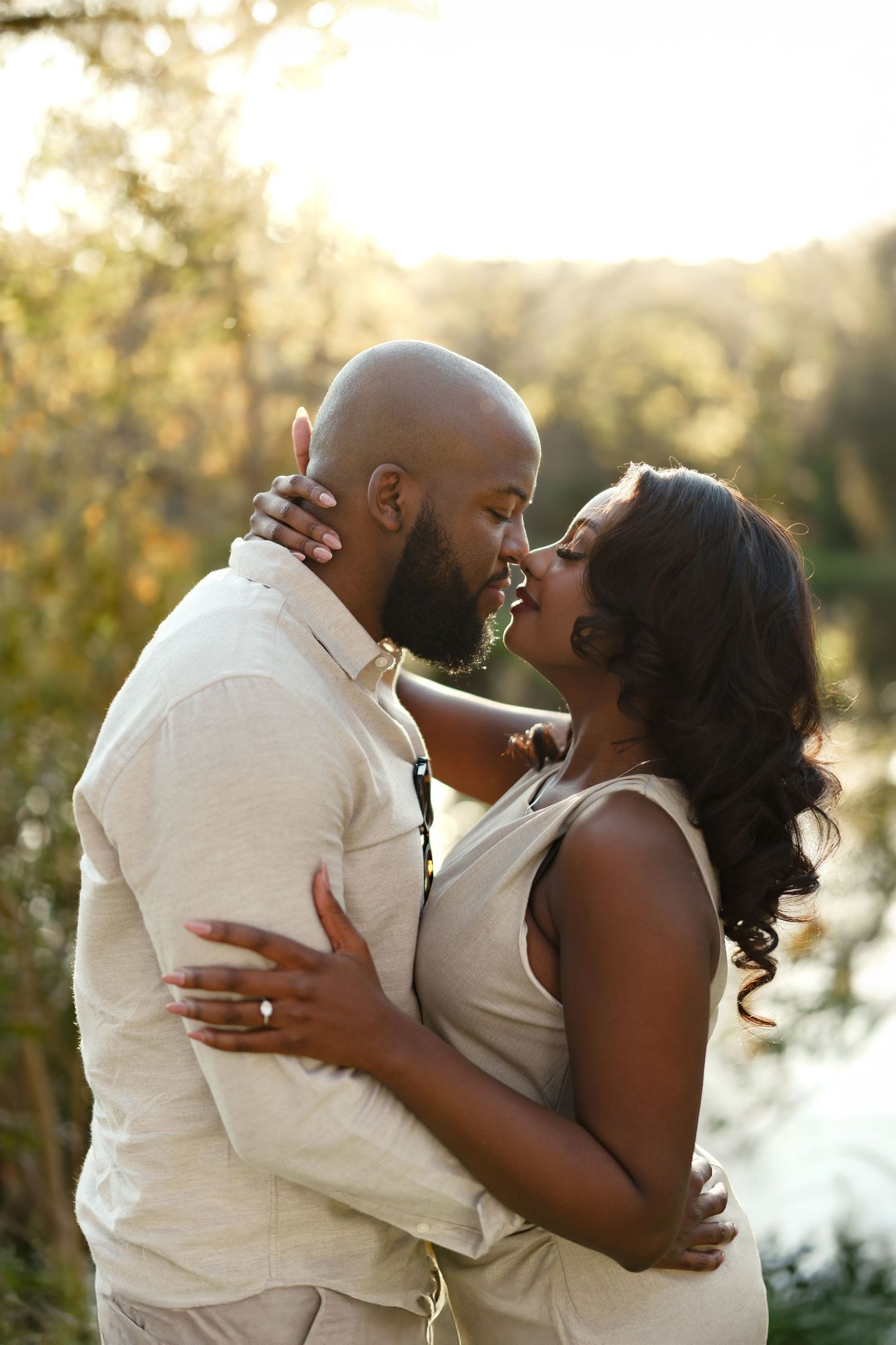 A romantic couple embraces outdoors during golden hour with soft sunlight filtering through trees in the background.