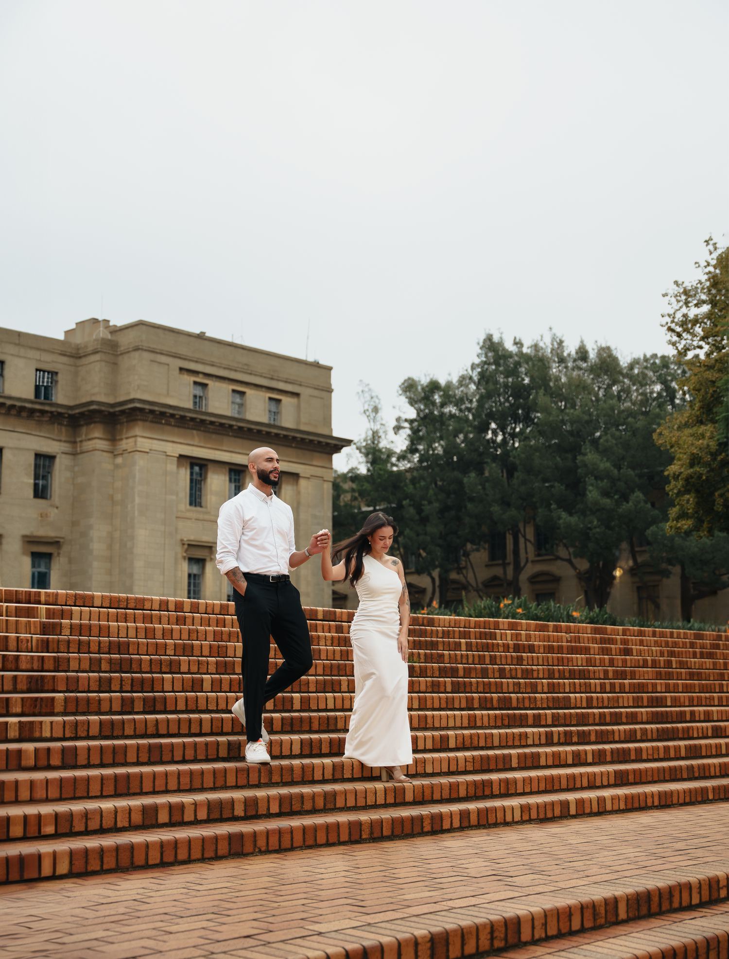 Couple in contrasting white and black outfits pose playfully on grand brick stairs of historic building.