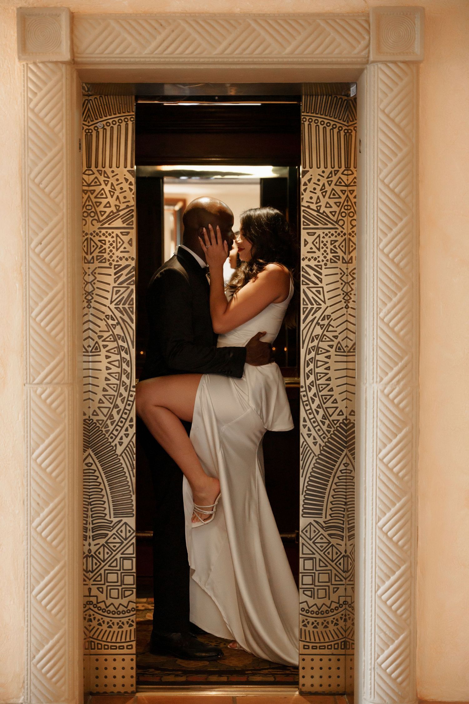 Romantic couple embraces in an ornate elevator doorway with the bride lifting her leg during a wedding photoshoot.
