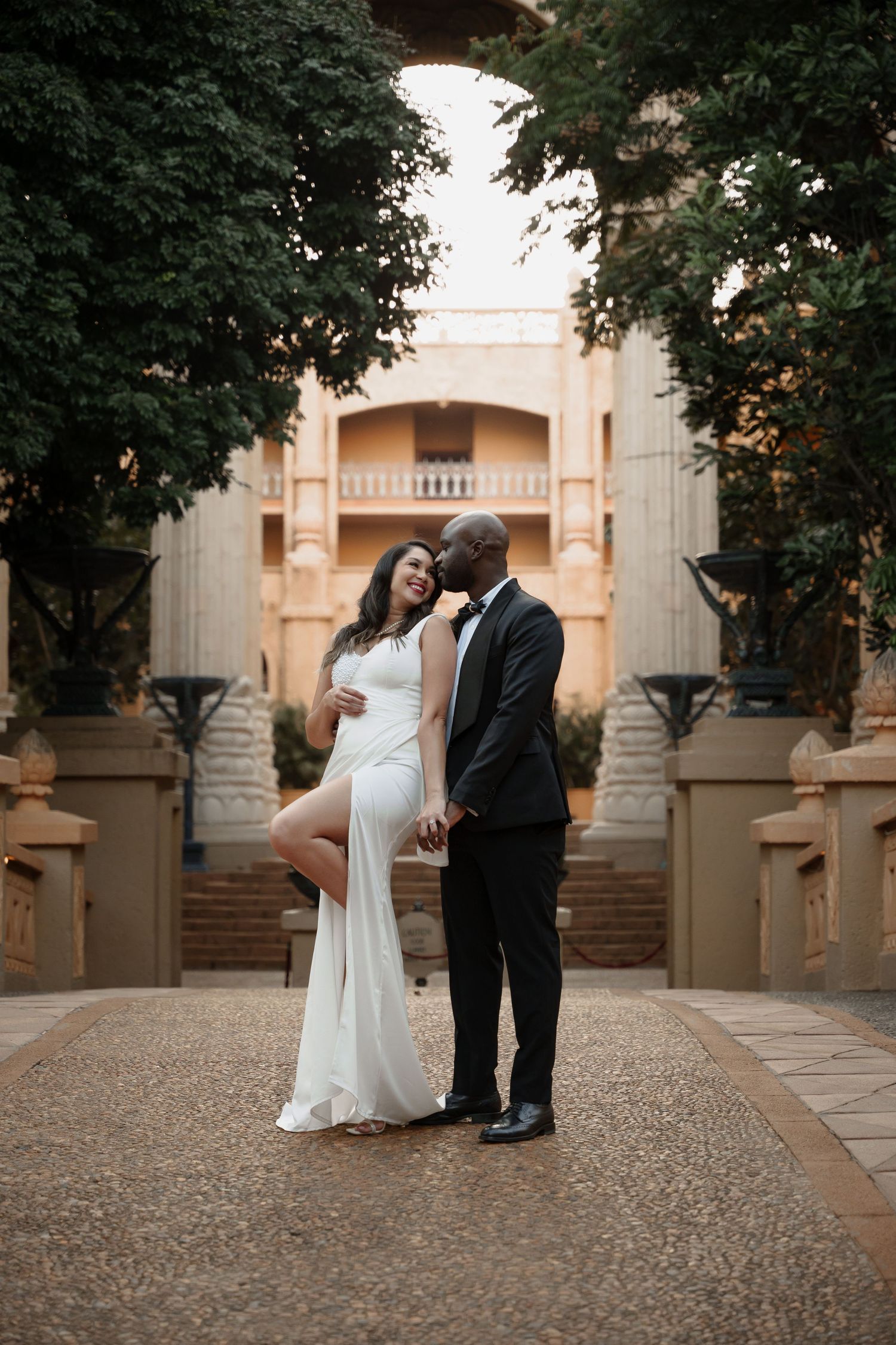 A romantic wedding couple poses on a grand architectural pathway framed by trees.