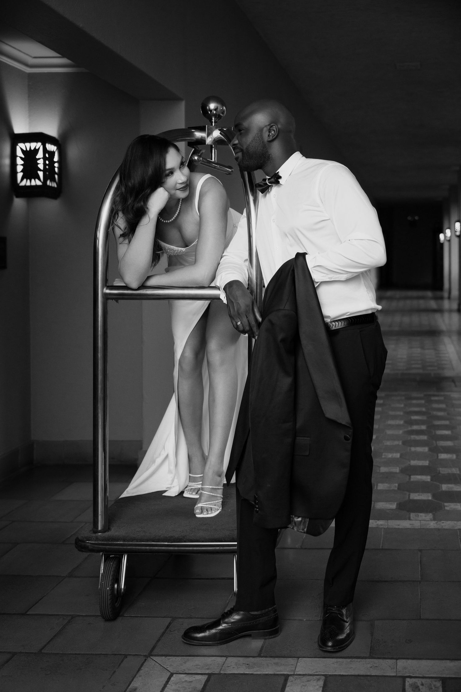 Creative wedding photo of couple posing with hotel luggage cart in a hallway.