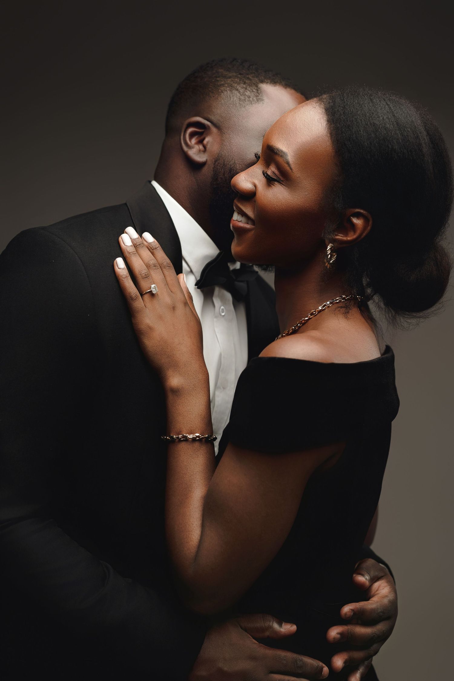 Elegant couple in formal black attire share an intimate moment in dramatic studio lighting.