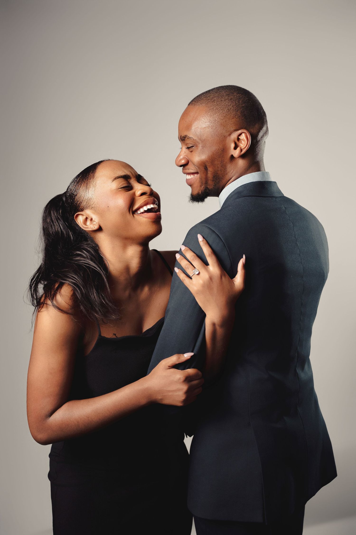 A joyful couple in formal black attire share an intimate laughing moment against a neutral background.