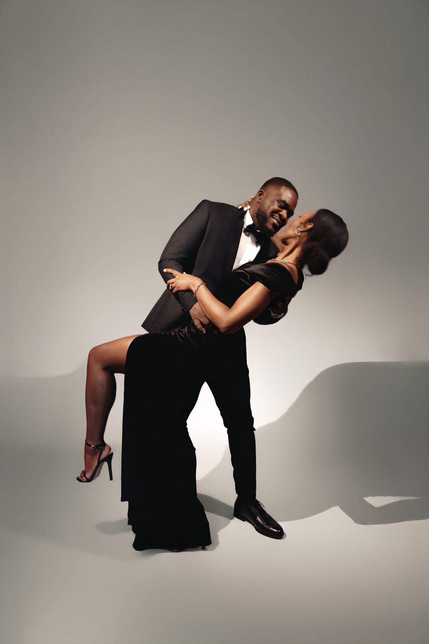 An elegant couple in formal attire performs a romantic dance pose on a minimalist gray background.