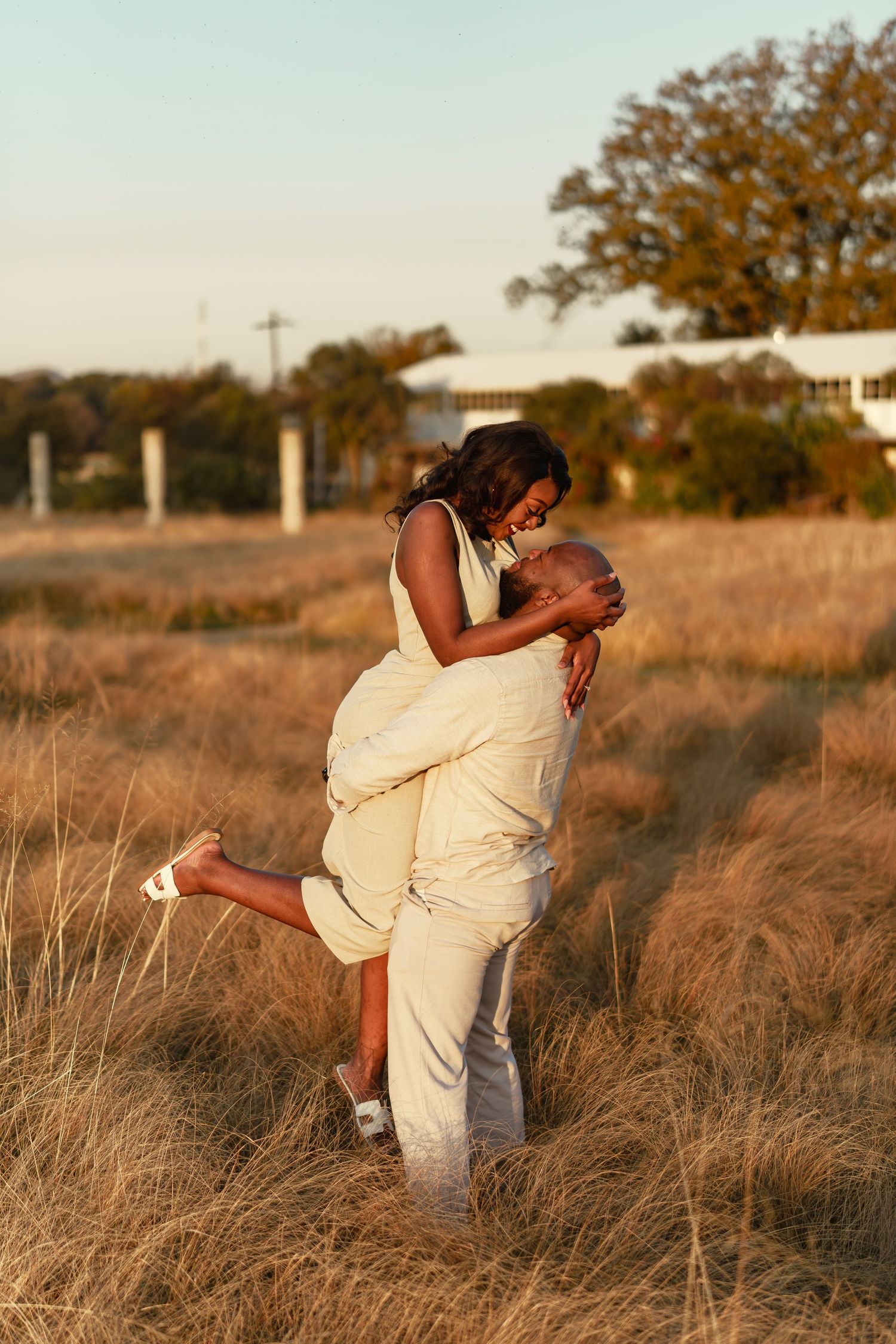 Romantic couple embracing in a golden field at sunset with tall grass and autumn trees in the background.