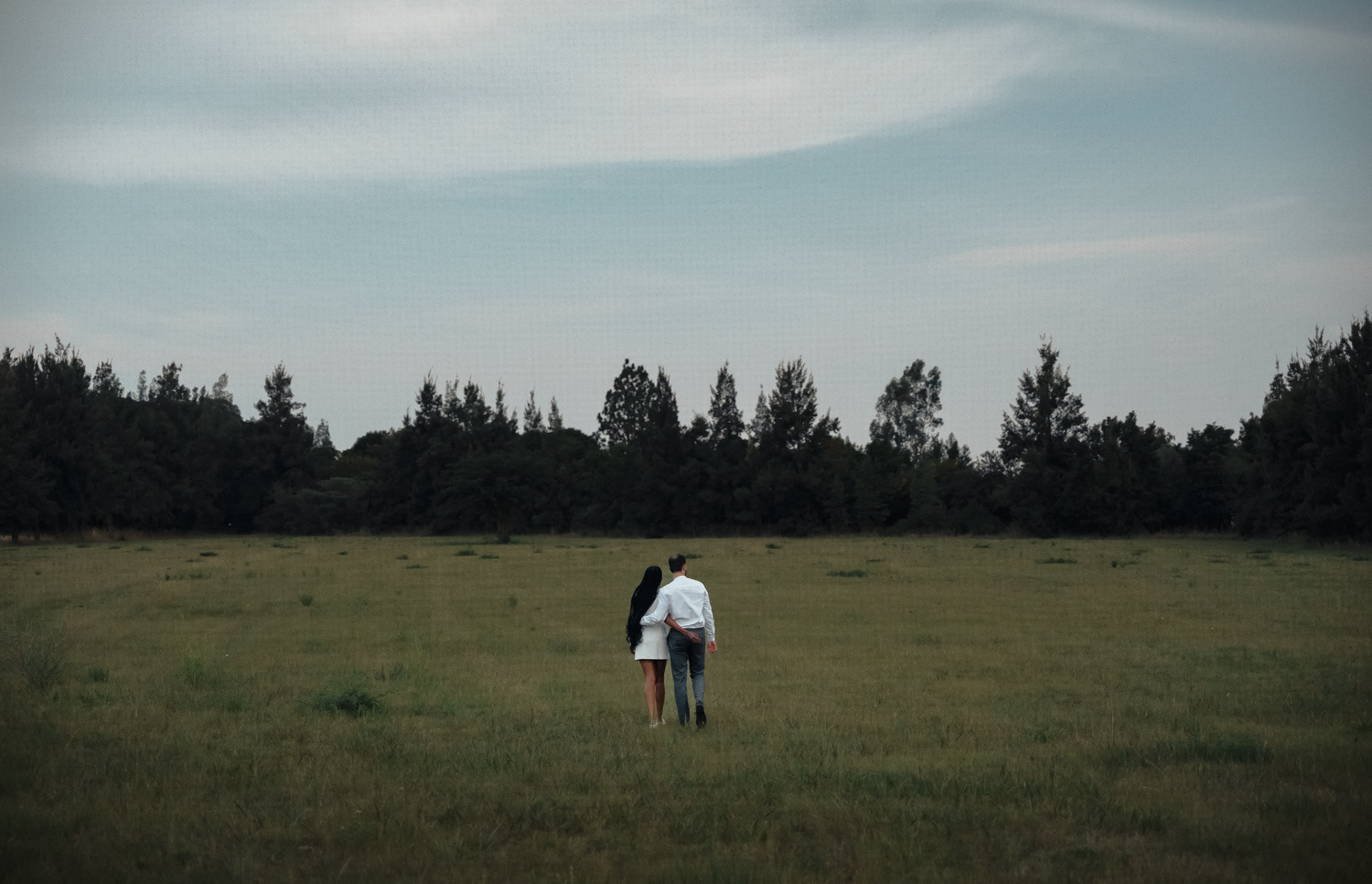 Silhouetted couple walking together in a meadow with pine trees and cloudy sky in the background.