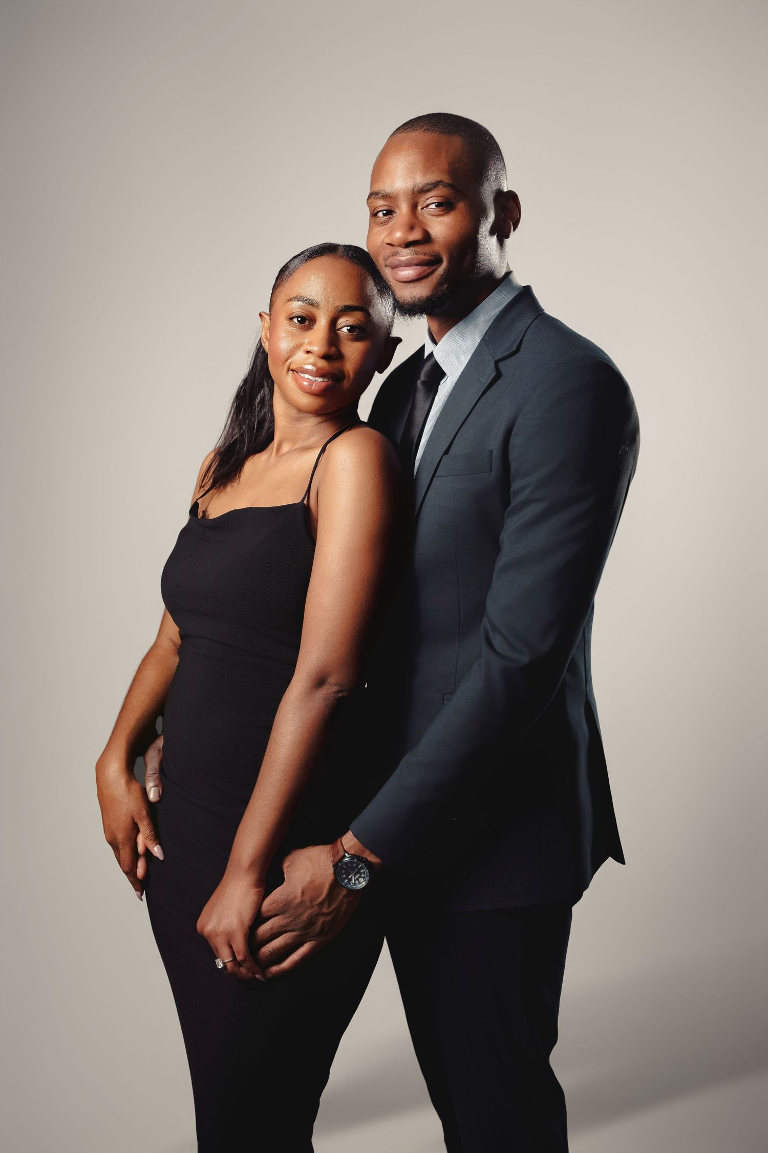 Elegant couple in formal black attire pose together against neutral backdrop for a romantic portrait session.
