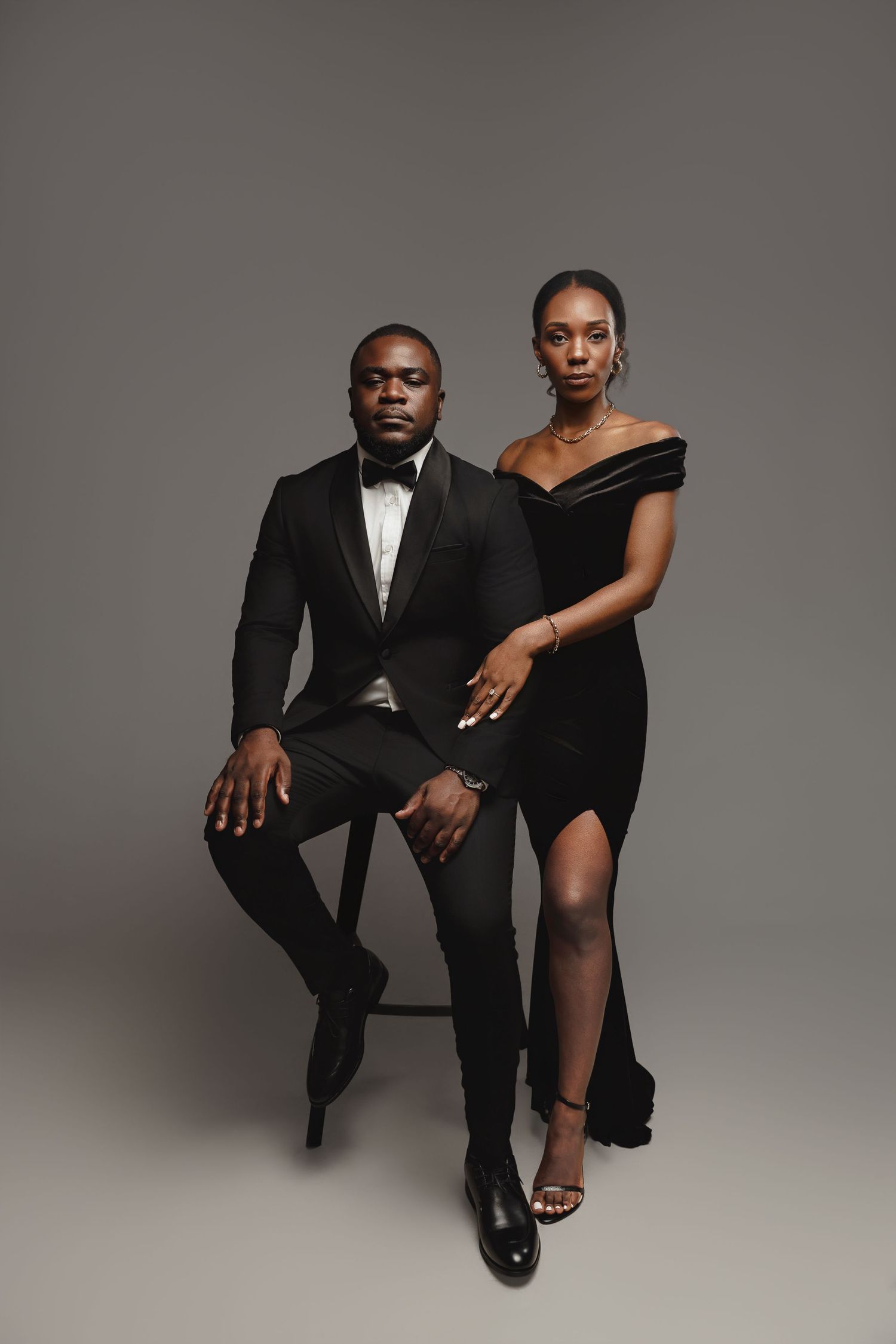 A stylish couple poses for artistic black and white fashion portraits in formal attire against a gray studio backdrop.