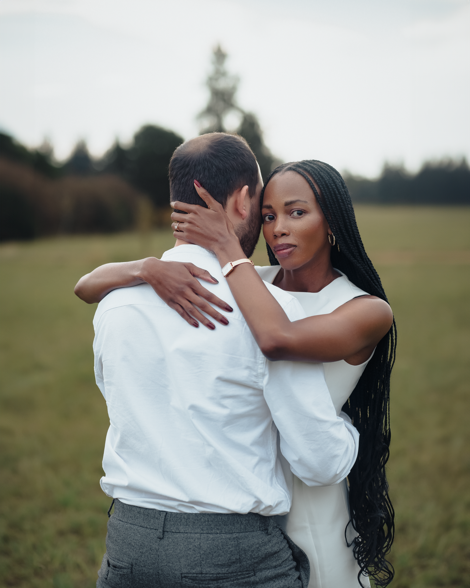 Romantic couple embracing in a field with forest backdrop during golden hour.