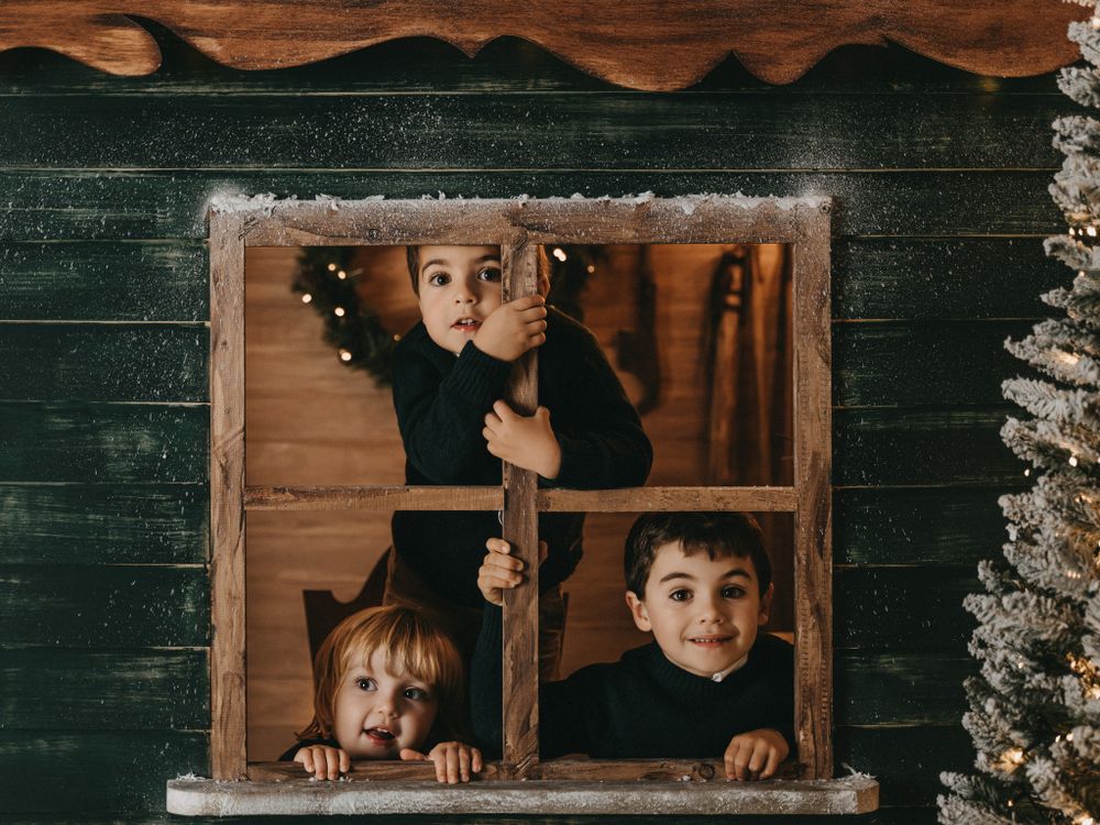 Three smiling faces peek through a wooden window frame with Christmas decorations visible in the background.
